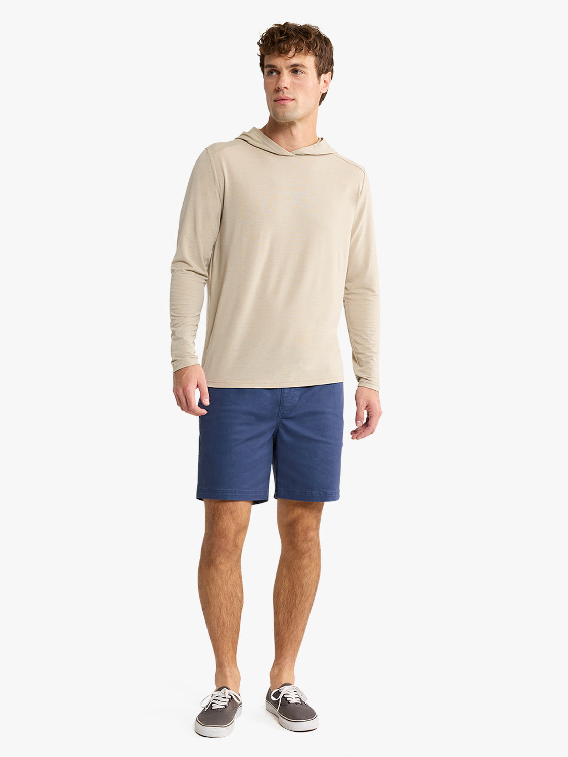 Man wearing a sand-colored long-sleeve hoodie with navy blue shorts and gray sneakers.