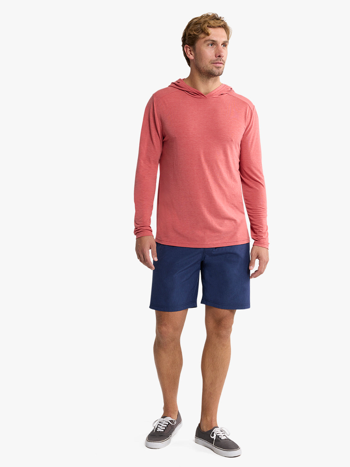Man wearing a mineral red hooded long-sleeve shirt with navy shorts and gray sneakers.