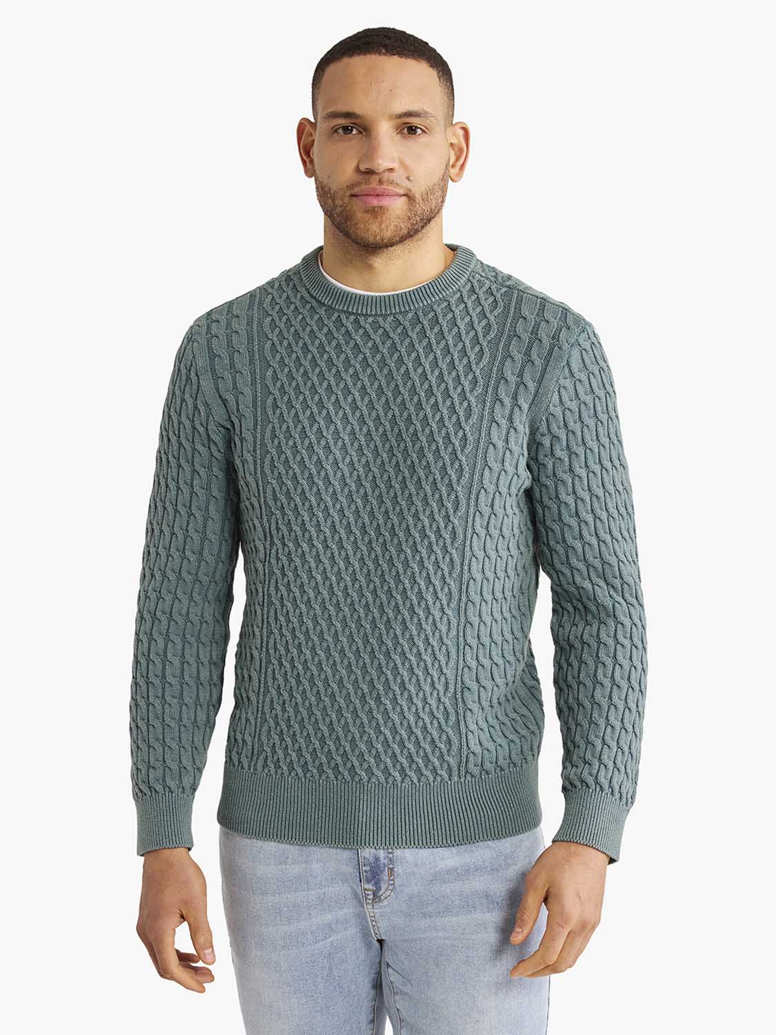 Man wearing a seapine green cable knit sweater with ribbed cuffs over light blue jeans.