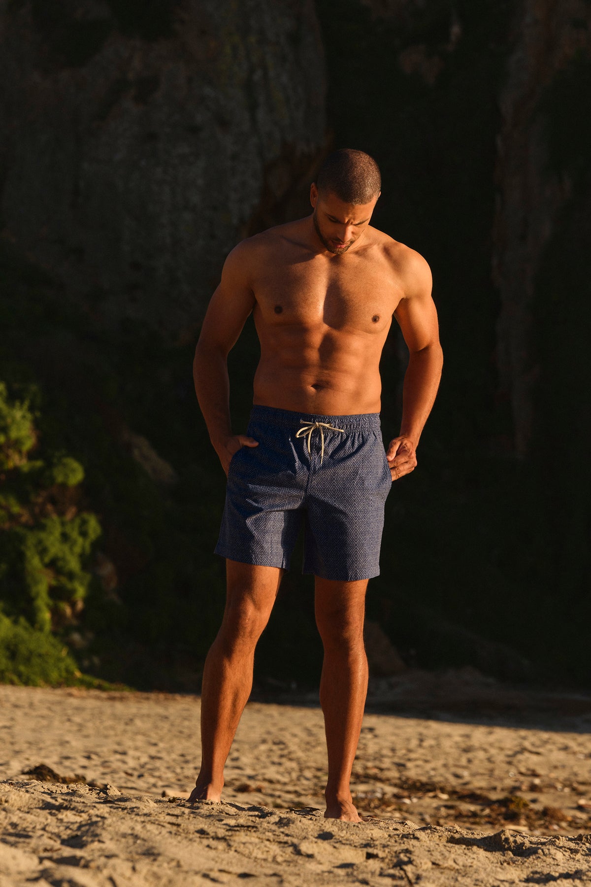 Man wearing navy shibori patterned swim shorts stands on sandy beach near dark foliage.