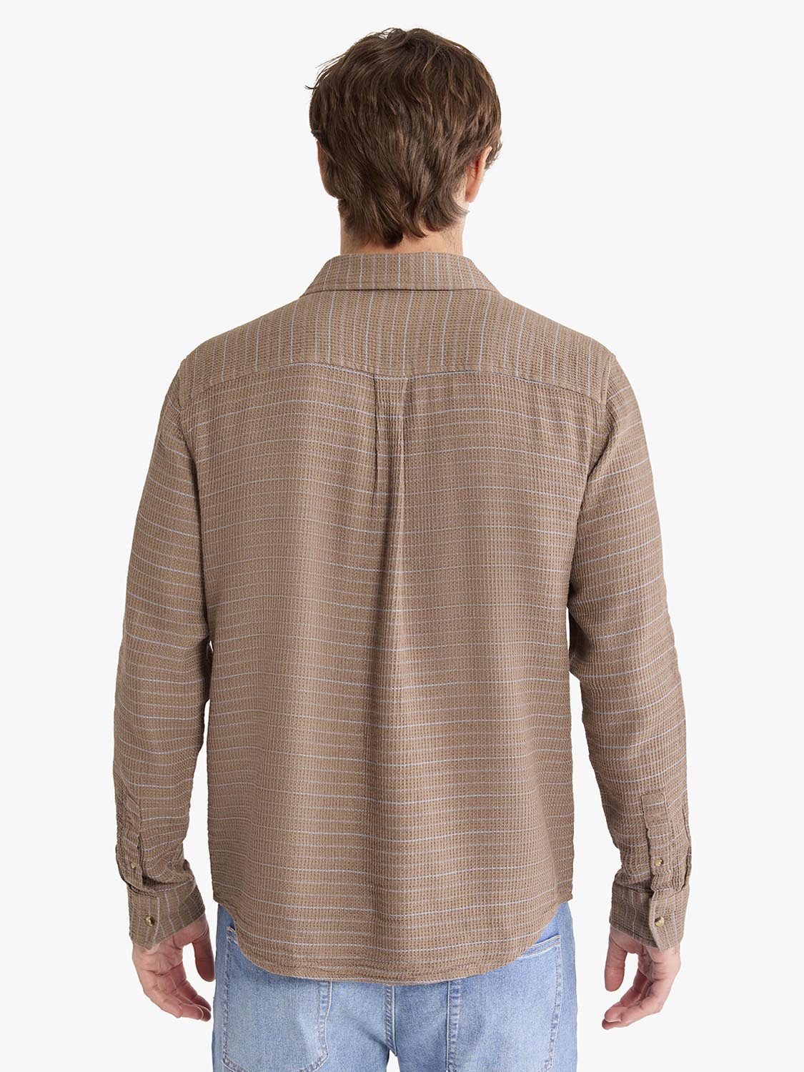 Man wearing a tan waffle stripe long sleeve button up shirt over blue jeans, shown from behind.