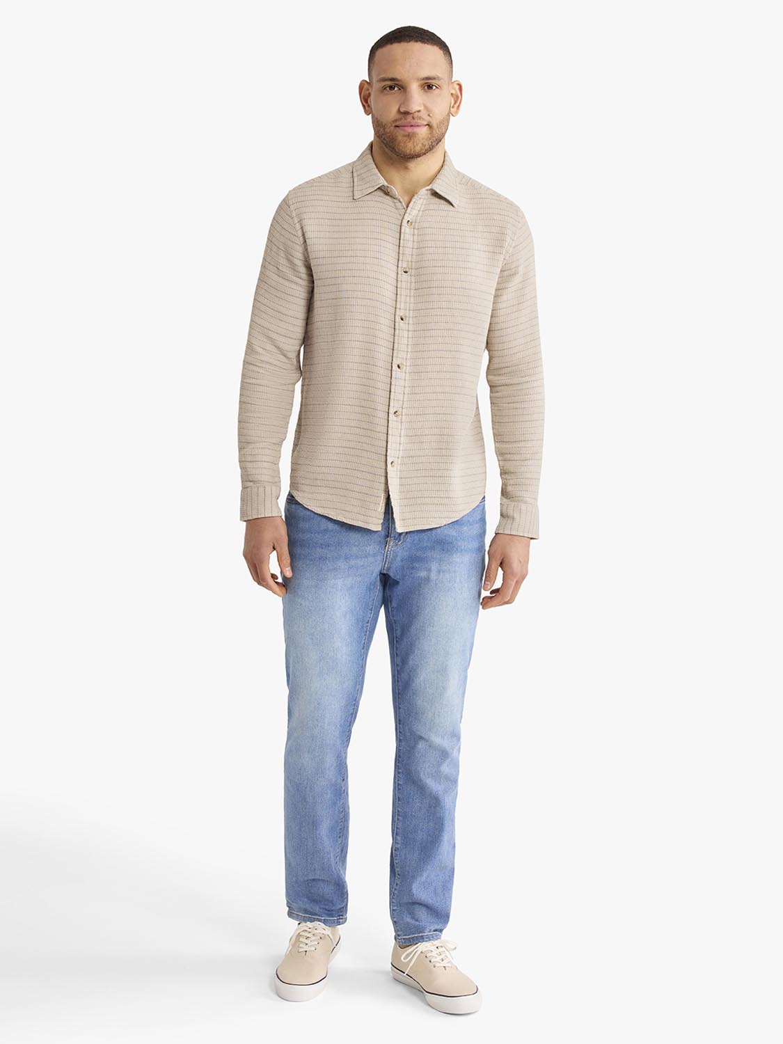 Man wearing a beige striped long sleeve button-up shirt with blue jeans and tan sneakers.