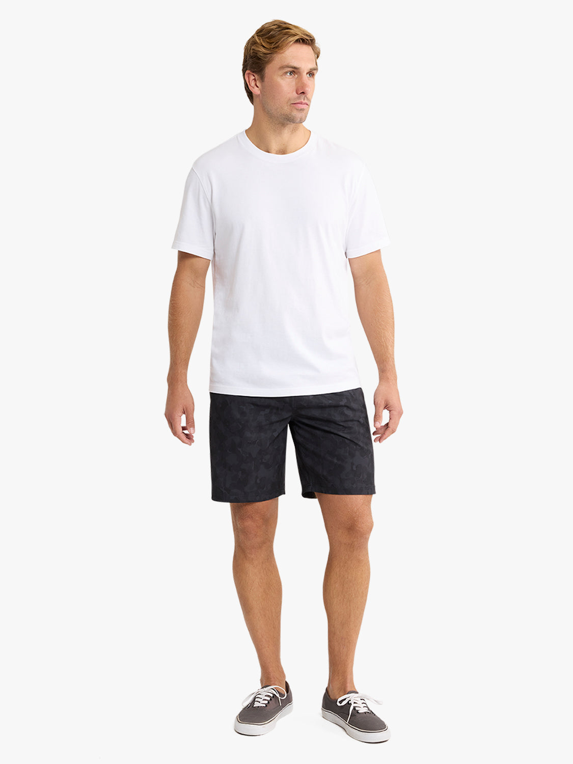 Man wearing a white crew-neck t-shirt and black camouflage shorts standing against a white background.
