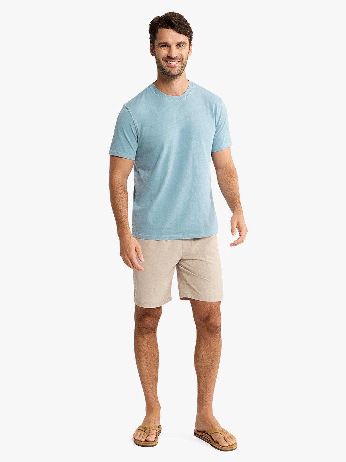 Man wearing light blue t-shirt and khaki shorts with tan sandals against white background.