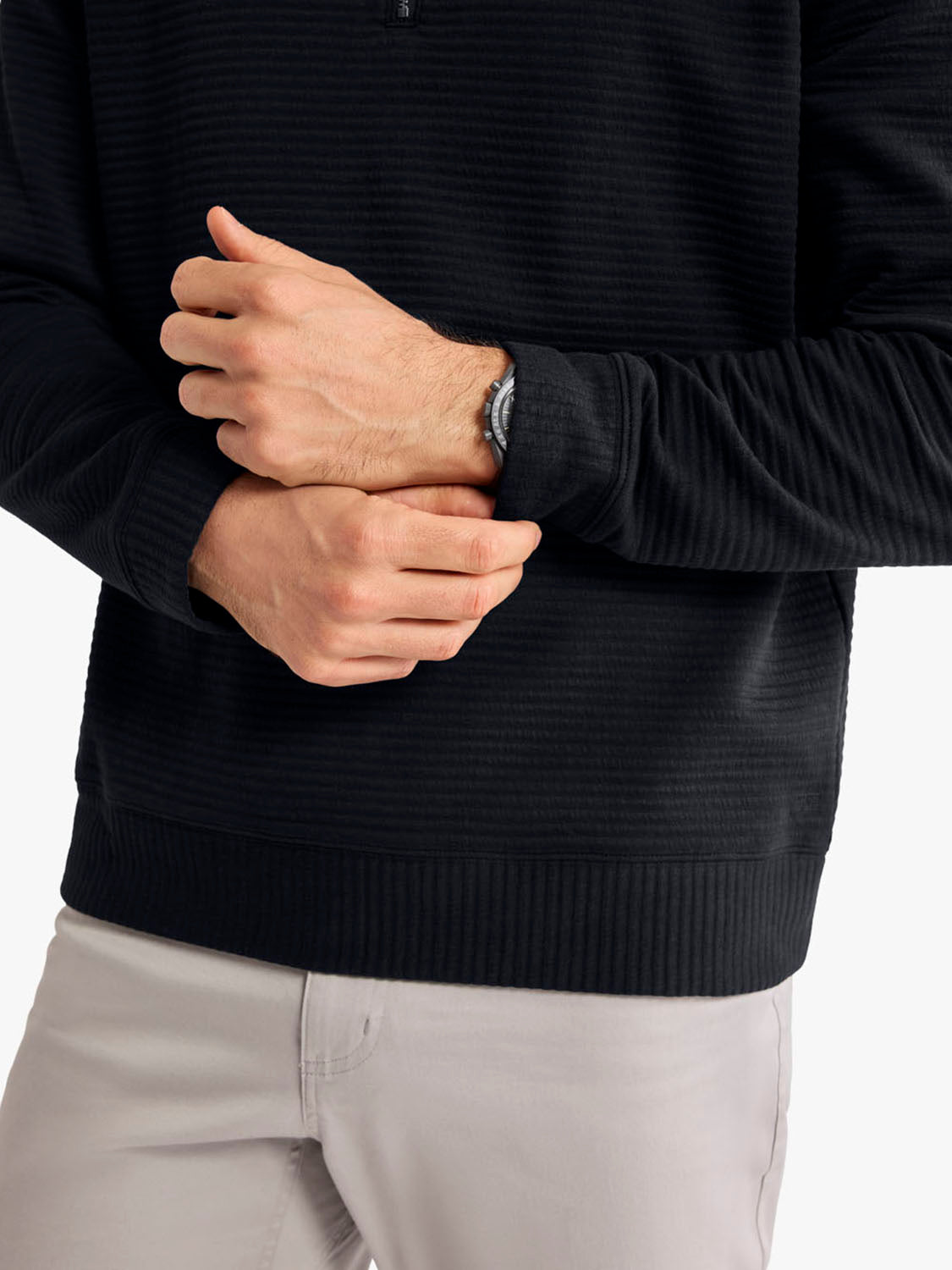 Man wearing a black ribbed quarter zip pullover with light gray pants, hands clasped at chest.