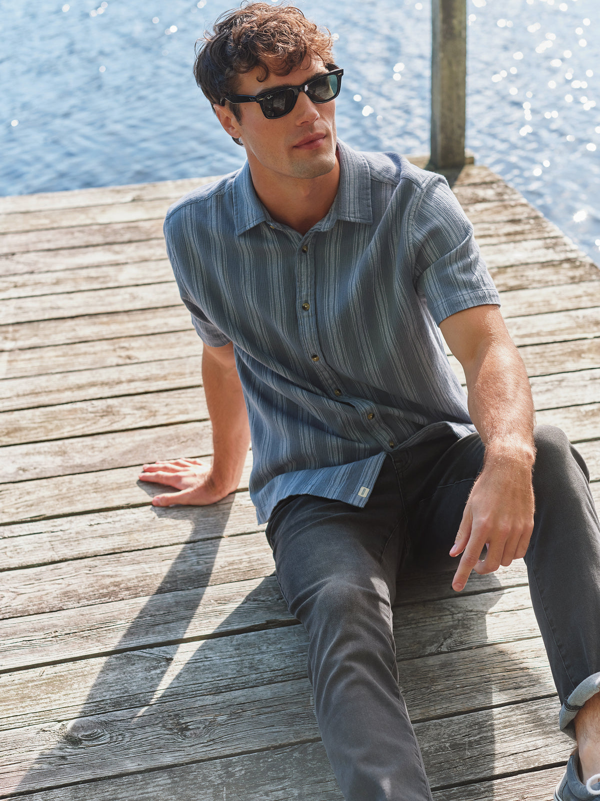 Man wearing blue striped short-sleeve shirt sitting on wooden dock beside water.