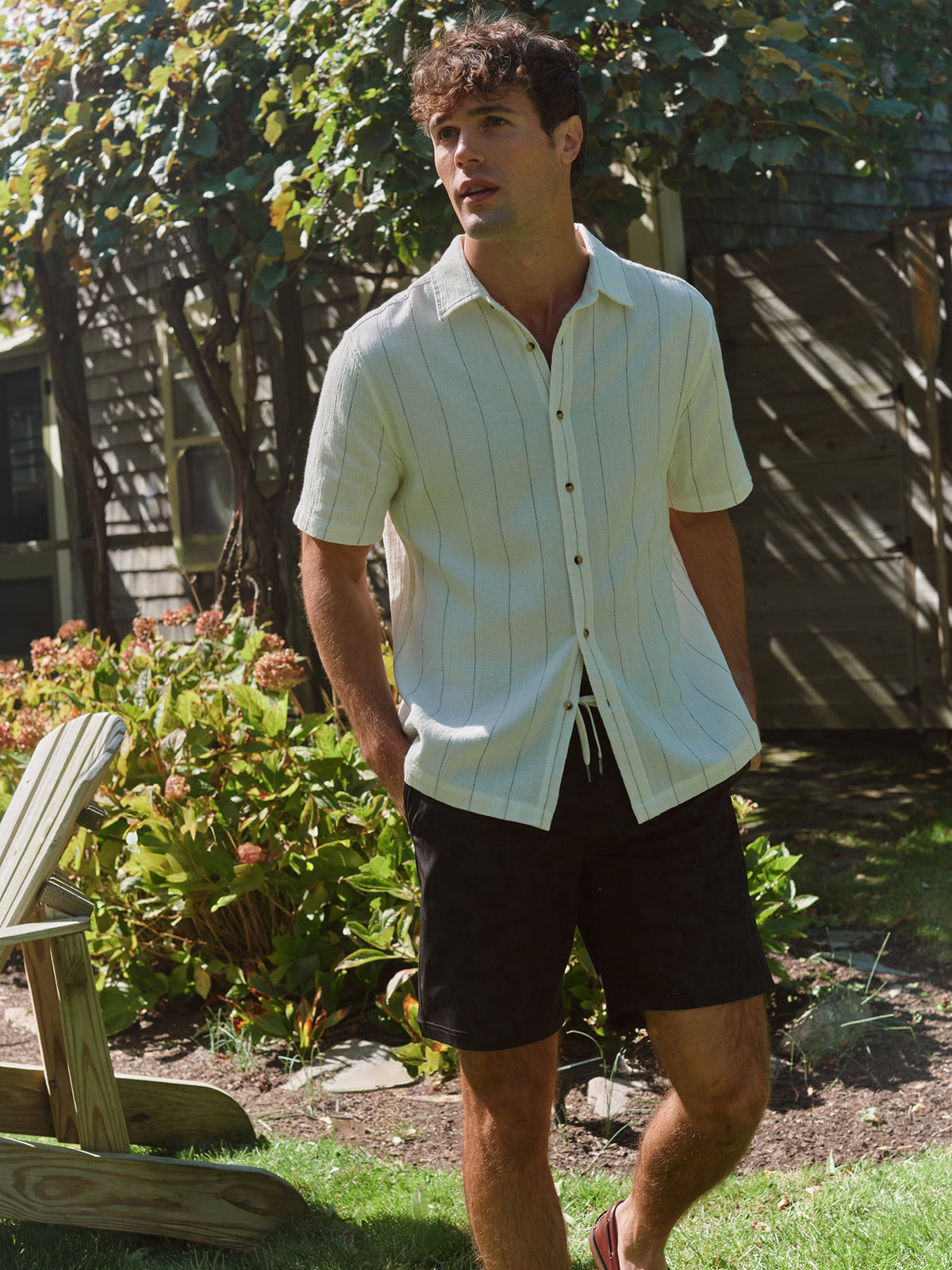 Man wearing a light yellow striped short-sleeve shirt and black shorts standing in a garden.