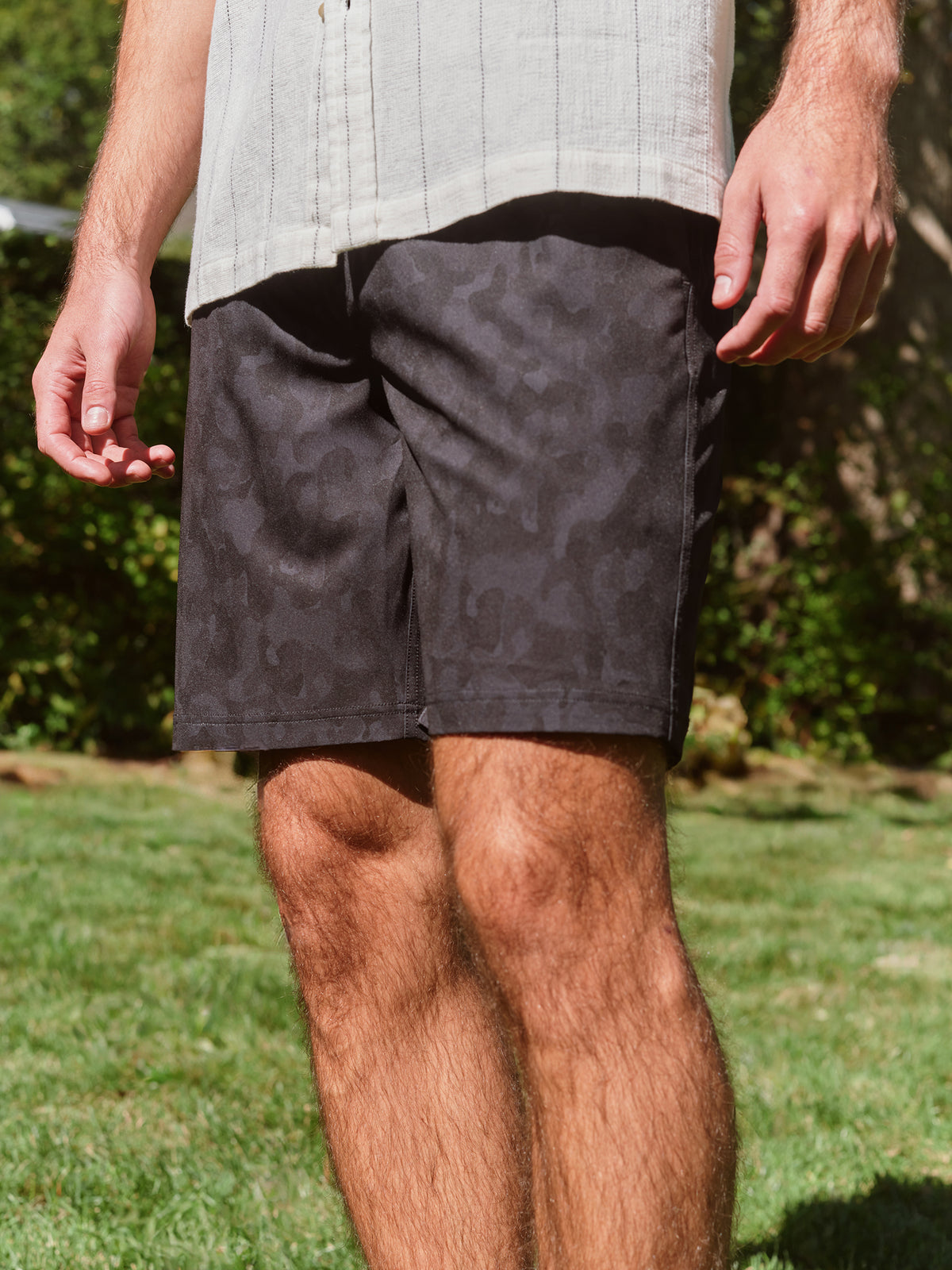 Man wearing black camouflage shorts and a light striped shirt standing outdoors.