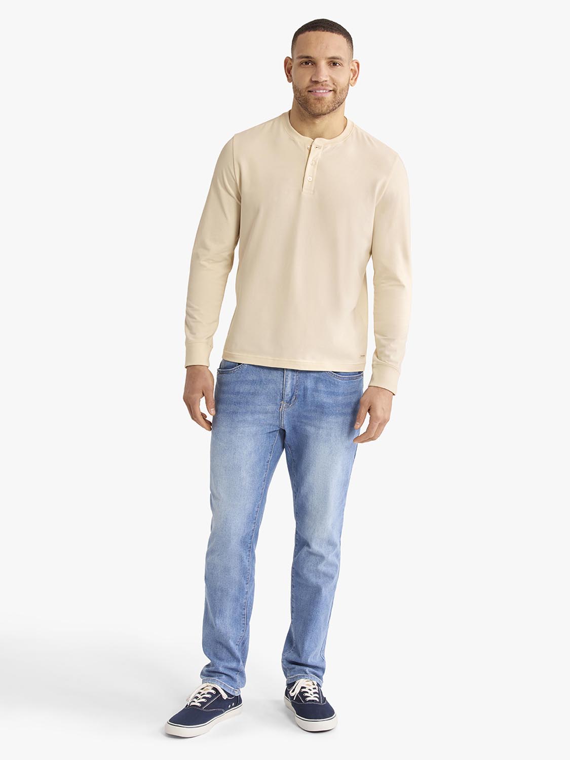 Man wearing a cream-colored long-sleeve henley shirt and blue jeans with navy sneakers.
