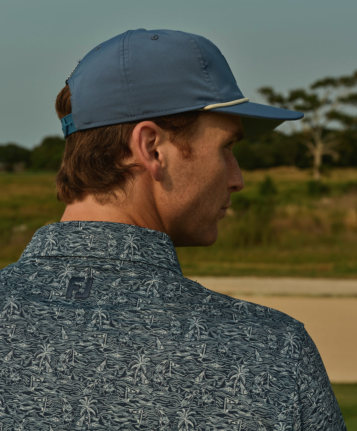 Man wearing blue baseball cap and tropical-patterned polo shirt standing outdoors on golf course.
