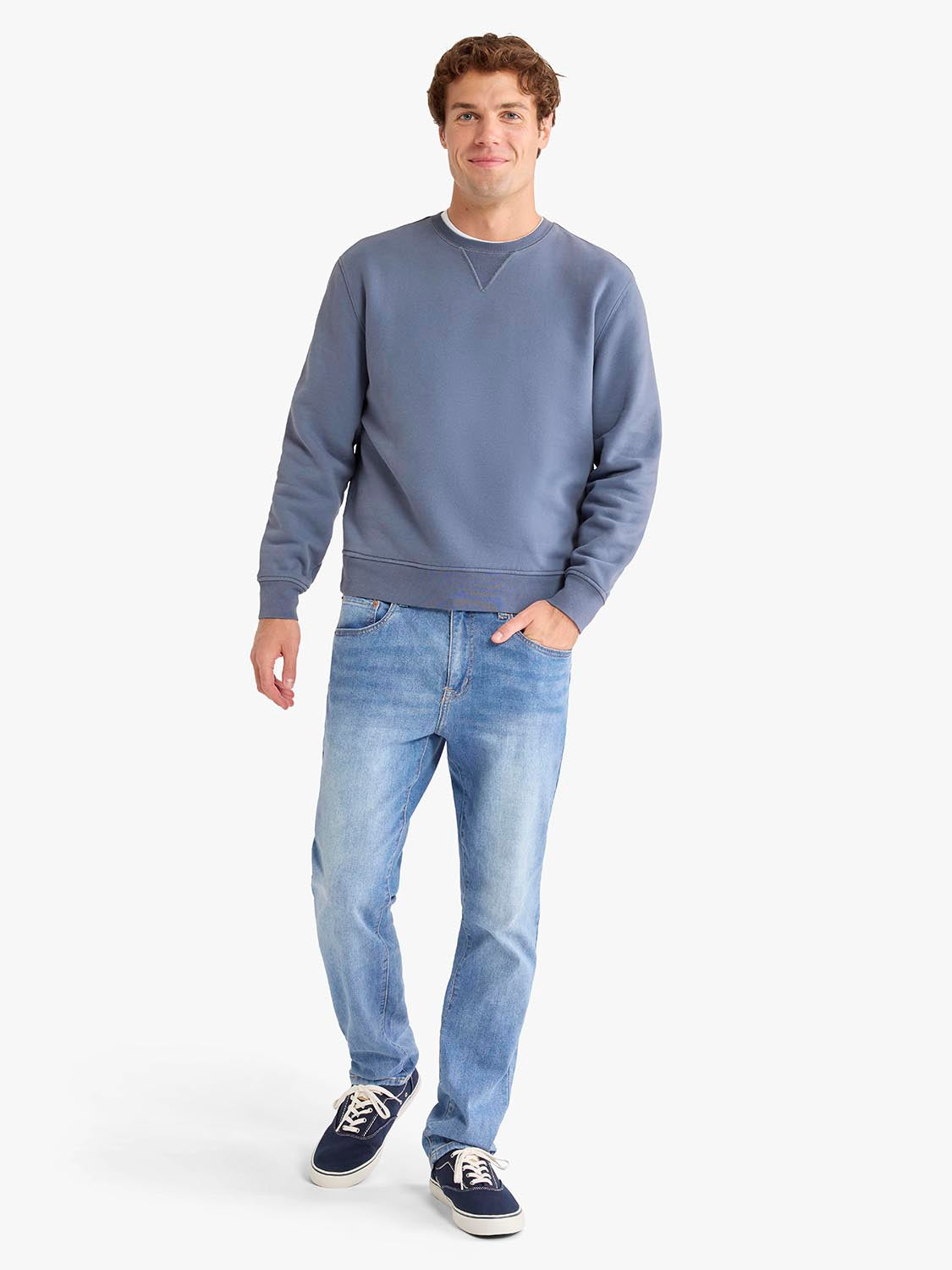 Man wearing a blue-gray crewneck sweatshirt with light blue jeans and navy sneakers.