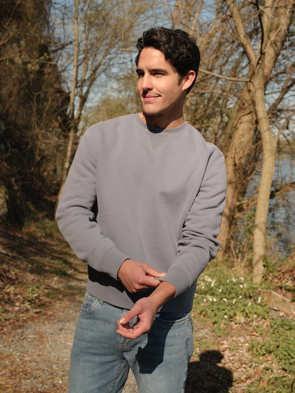 Man wearing a gray crewneck sweatshirt and light blue jeans standing outdoors near bare trees.