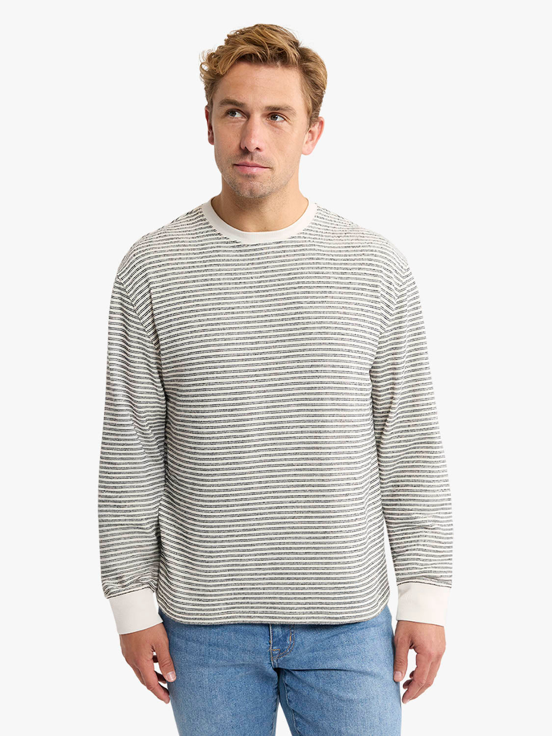 Man wearing a cream and gray horizontal striped crewneck sweatshirt with white cuffs and blue jeans.
