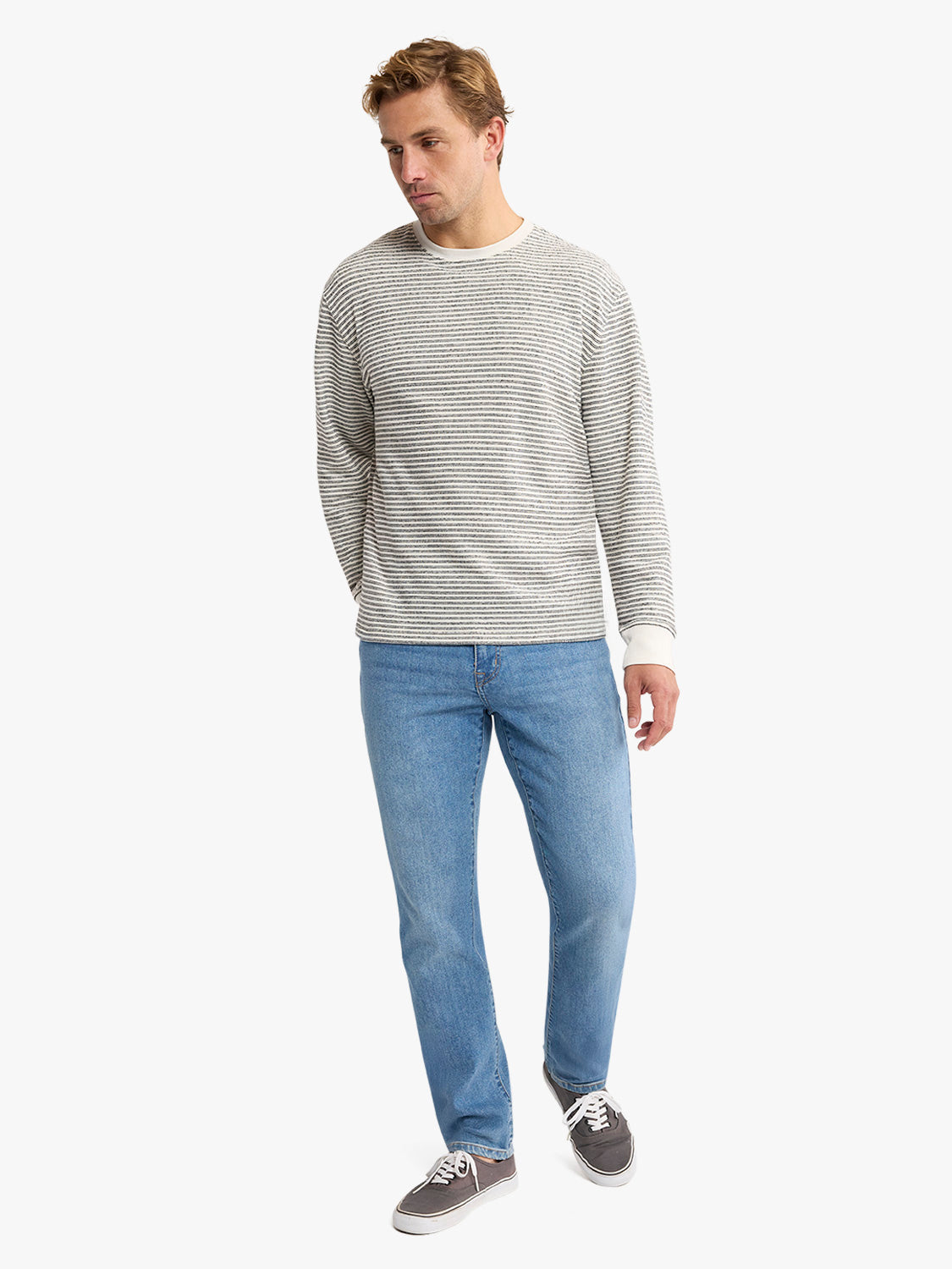 Man wearing a cream and gray striped crewneck sweatshirt with blue jeans and gray sneakers.