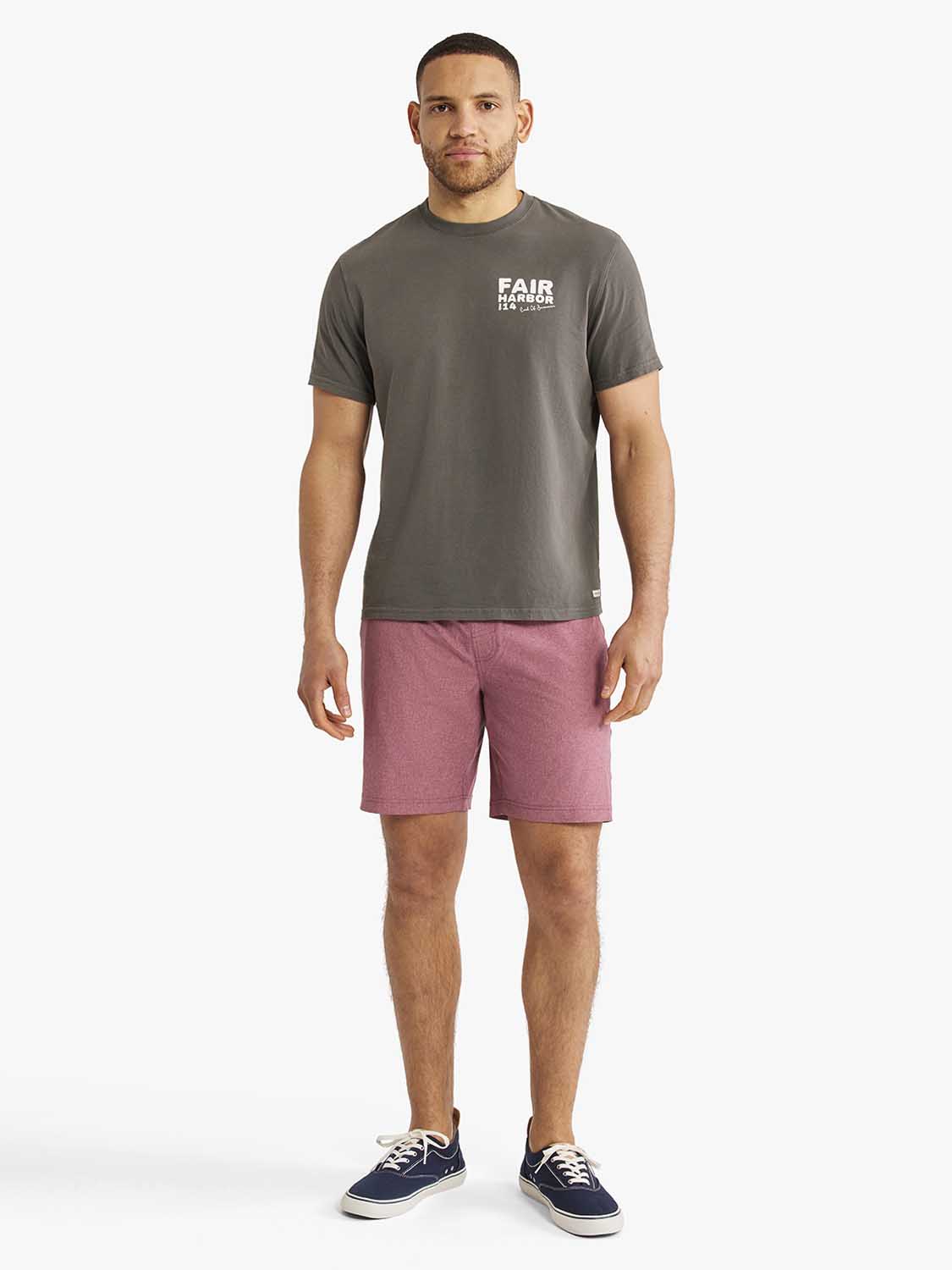Man wearing charcoal gray graphic tee with white text and mauve shorts with navy sneakers.