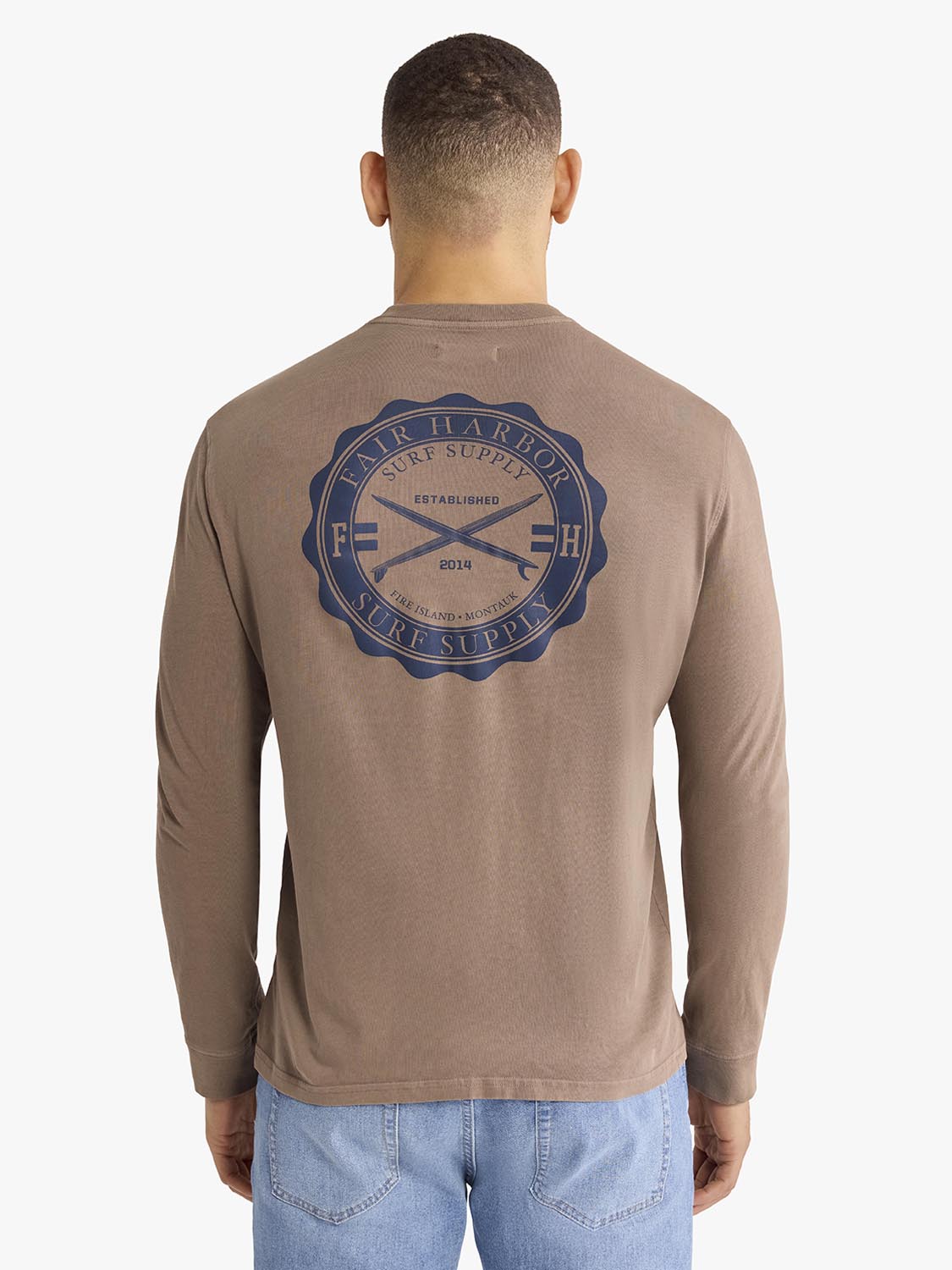 Man wearing taupe long sleeve tee with blue archival crest graphic on back, shown from behind.