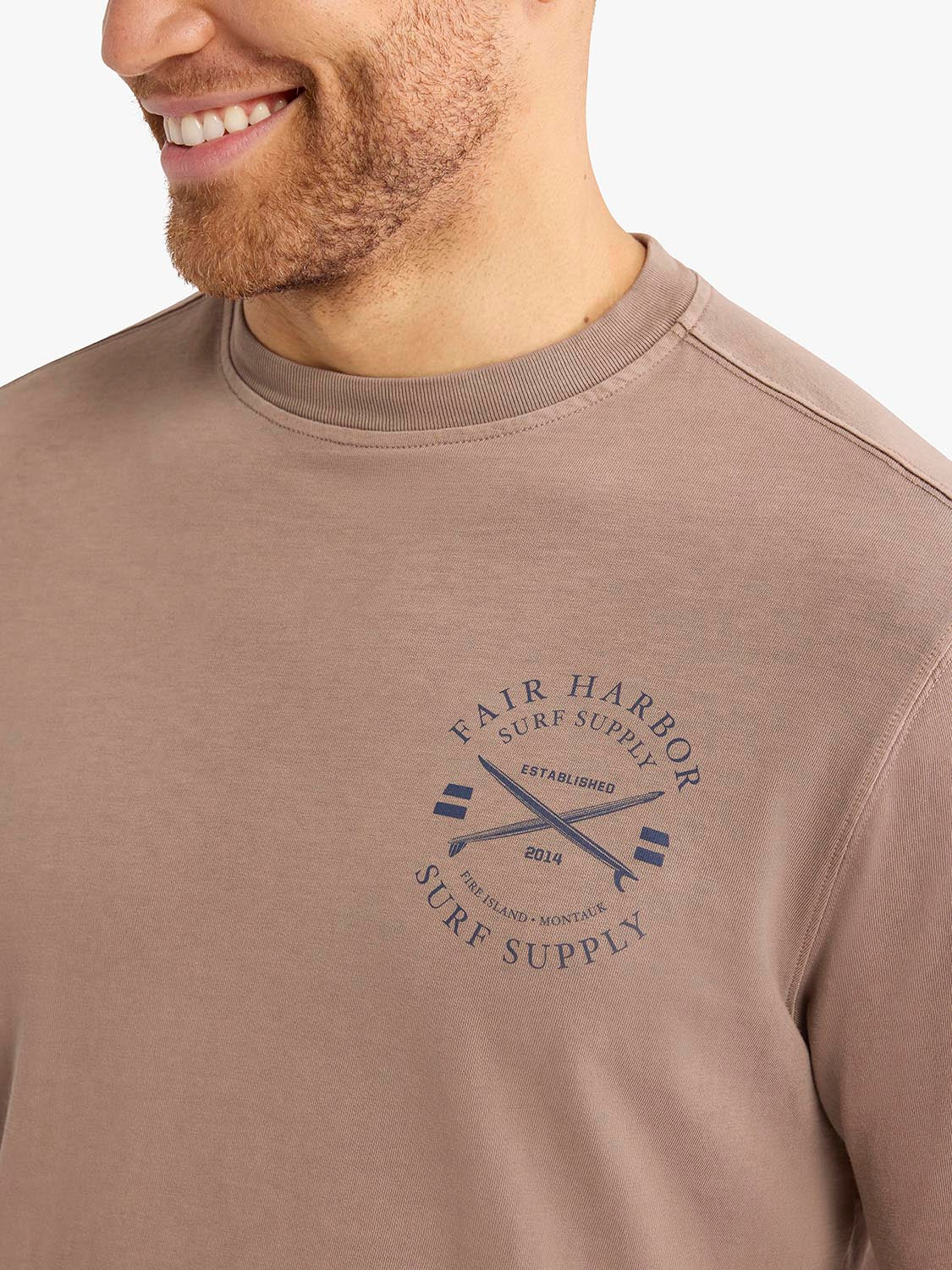 Man wearing a taupe long sleeve tee with a navy archival crest graphic on the chest.