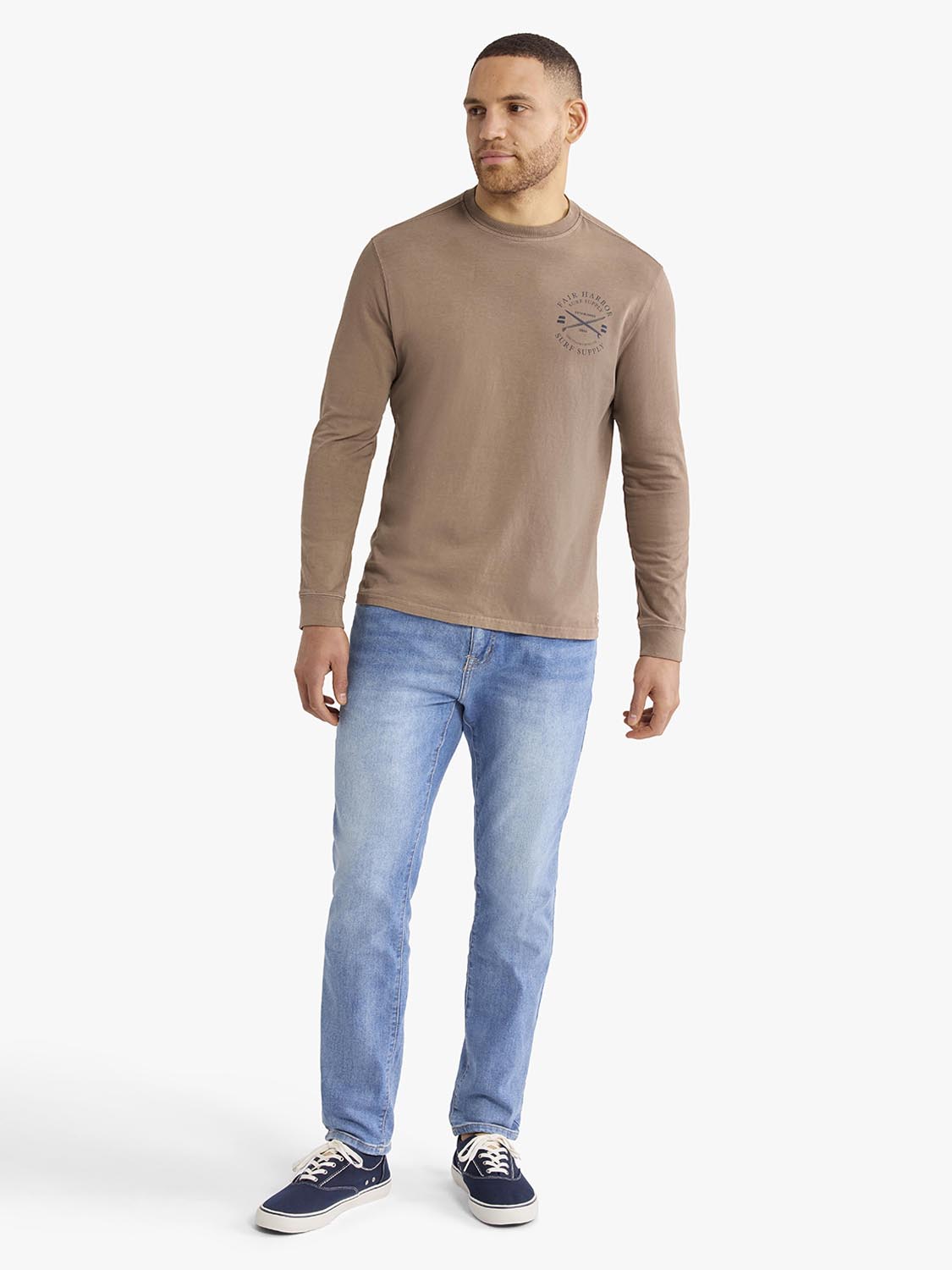 Man wearing a taupe long-sleeve graphic tee with archival crest design on chest.