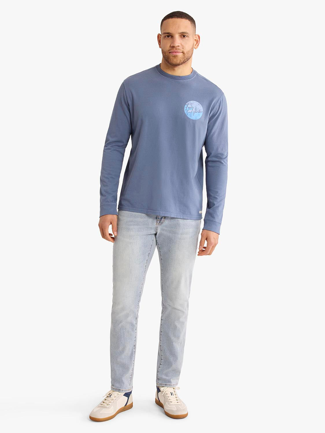 Man wearing a blue-grey long sleeve graphic tee with a circular chest design and light wash jeans.