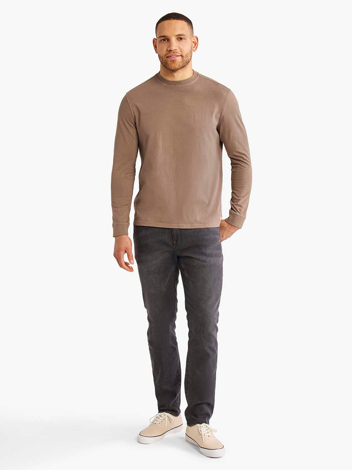 Man wearing a sandbar-colored long sleeve crew neck tee with dark jeans and beige sneakers.