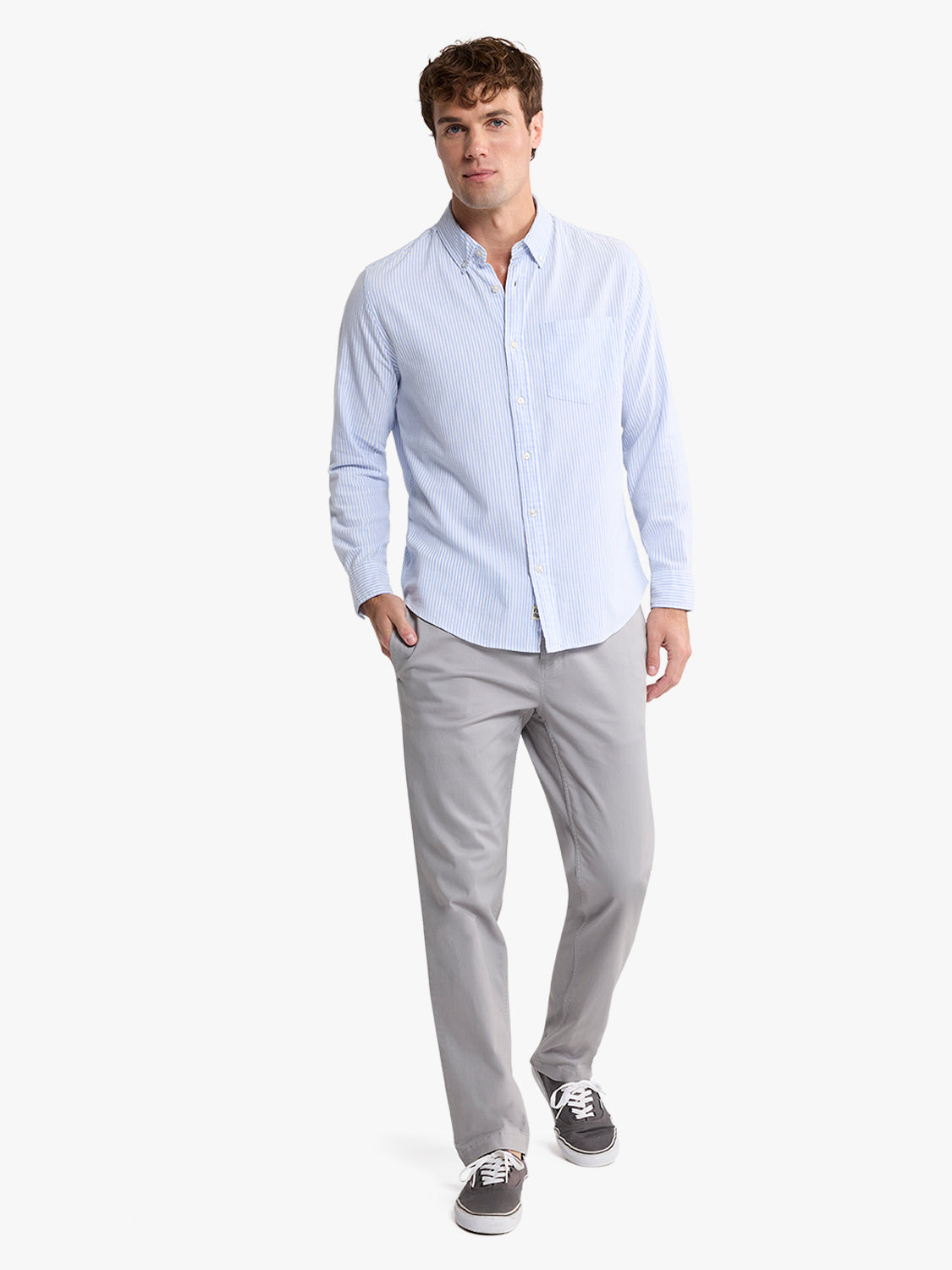 Man wearing light blue striped oxford shirt with gray pants and dark sneakers.