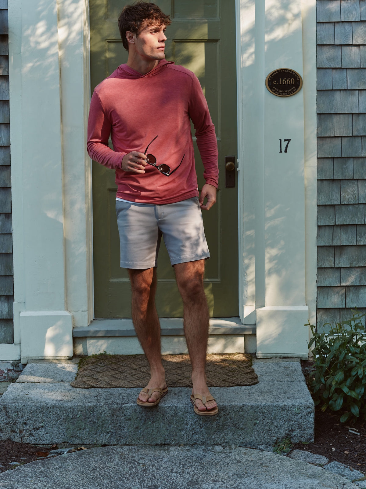 Man wearing a burgundy hoodie and light blue shorts standing in front of a coastal house entrance.