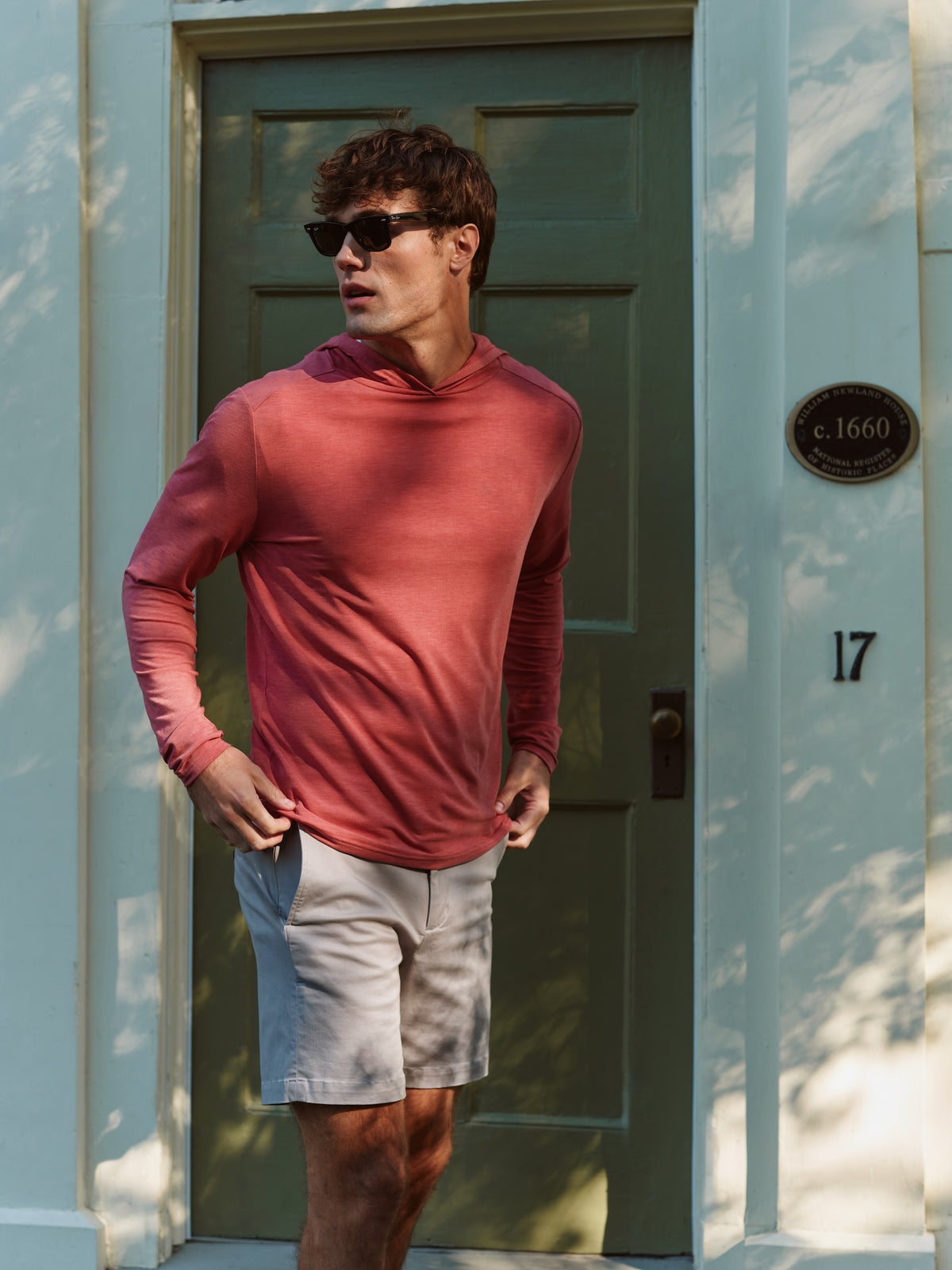 Man wearing a burgundy long-sleeve hoodie with light gray shorts and black sunglasses standing in a doorway.