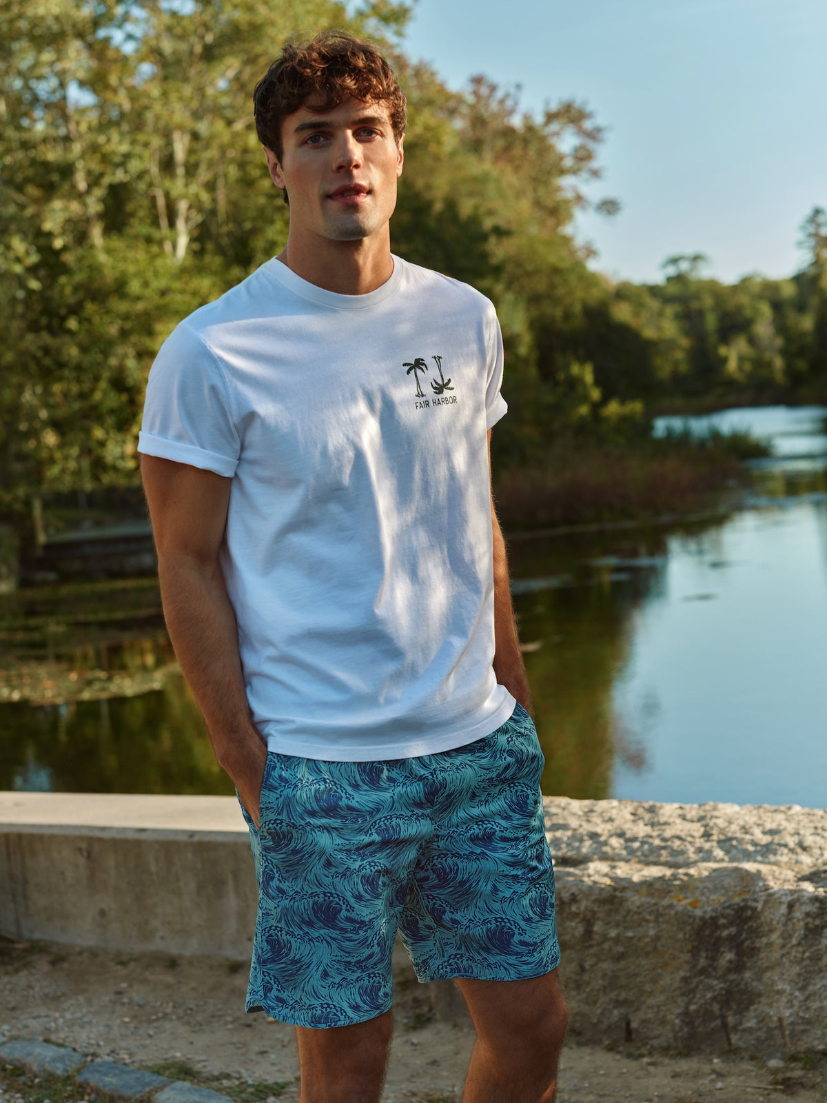 Man wearing white graphic t-shirt with palm tree design standing by a river.