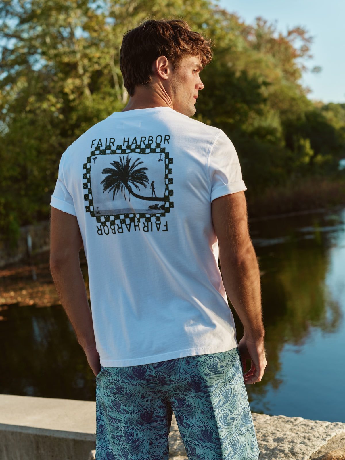 Man wearing white short-sleeve graphic tee with Fair Harbor and checkered palm tree design on back.
