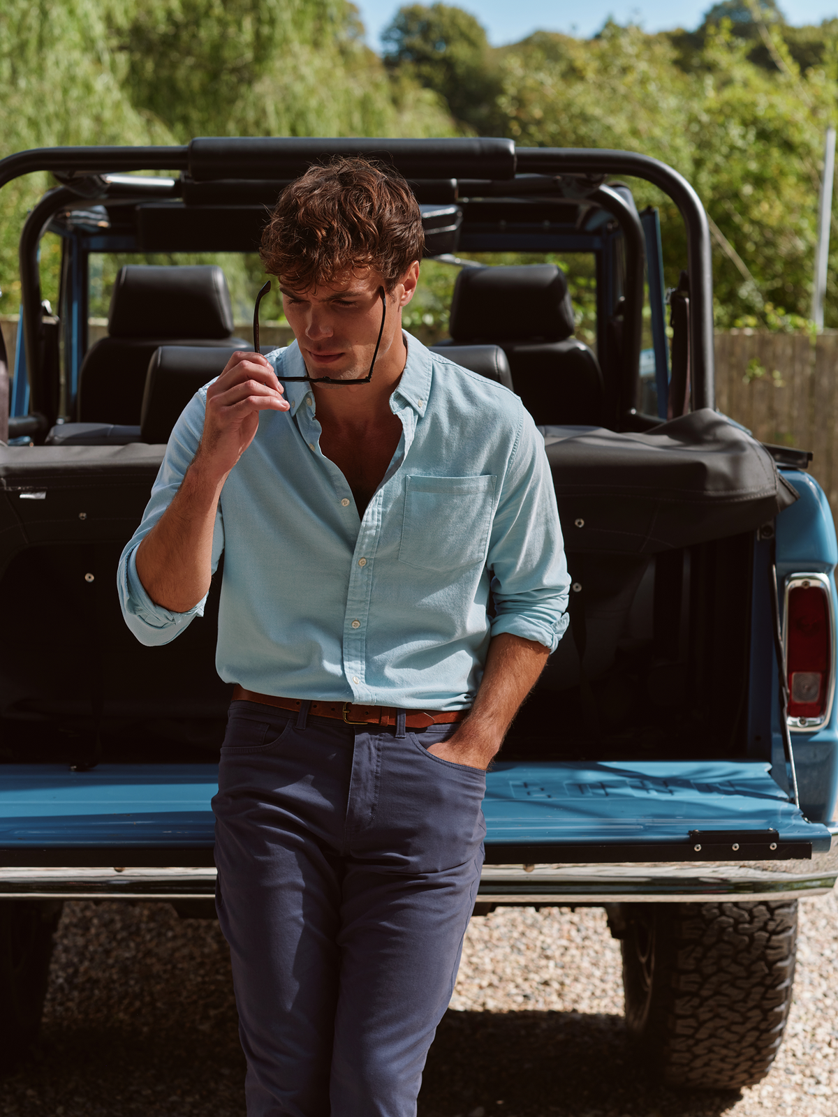 Man wearing a light blue striped oxford shirt and dark navy pants standing in front of a truck.