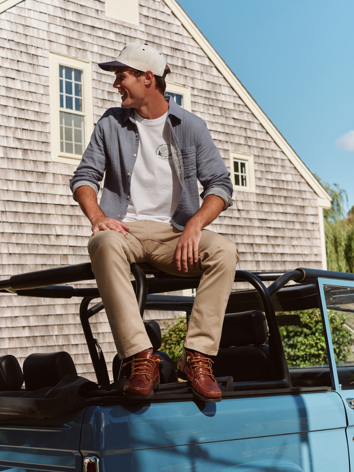 Man wearing gray button-up shirt over white t-shirt, sitting on truck bed beside coastal house.