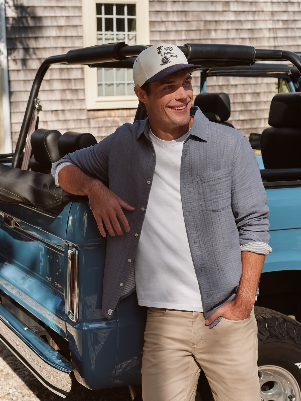 Man wearing a white t-shirt under a gray button-up shirt, leaning against a blue convertible truck.