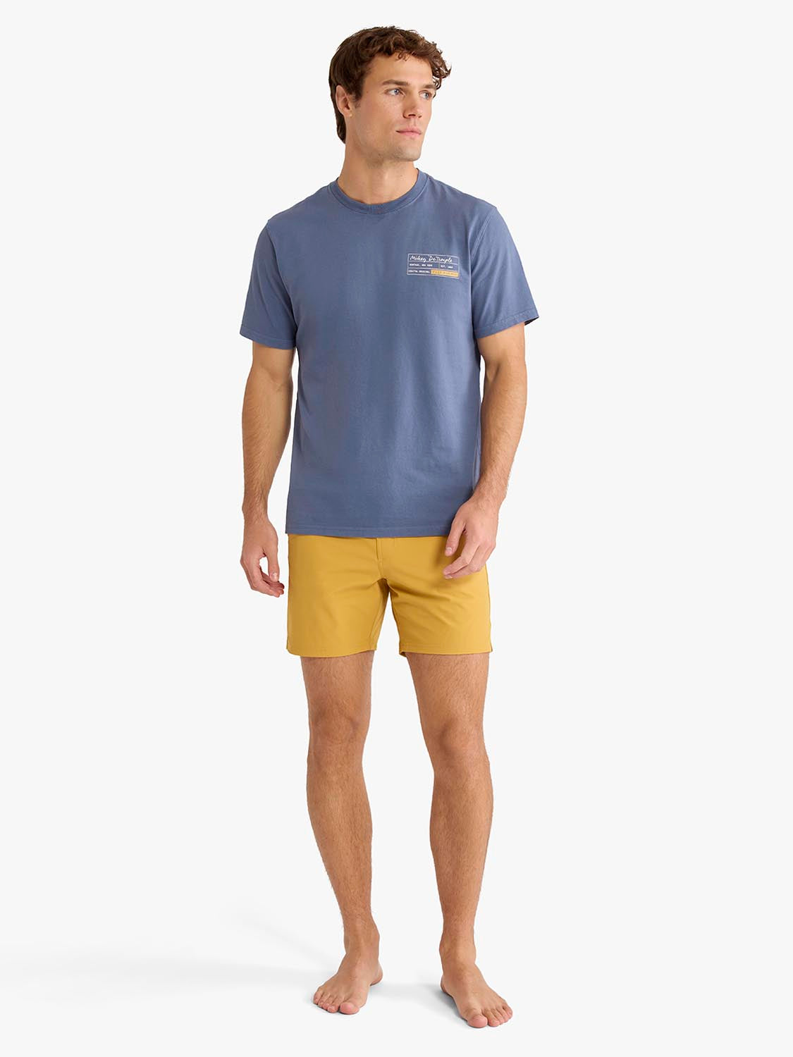 Man wearing gold board shorts with a blue t-shirt, standing against white background.