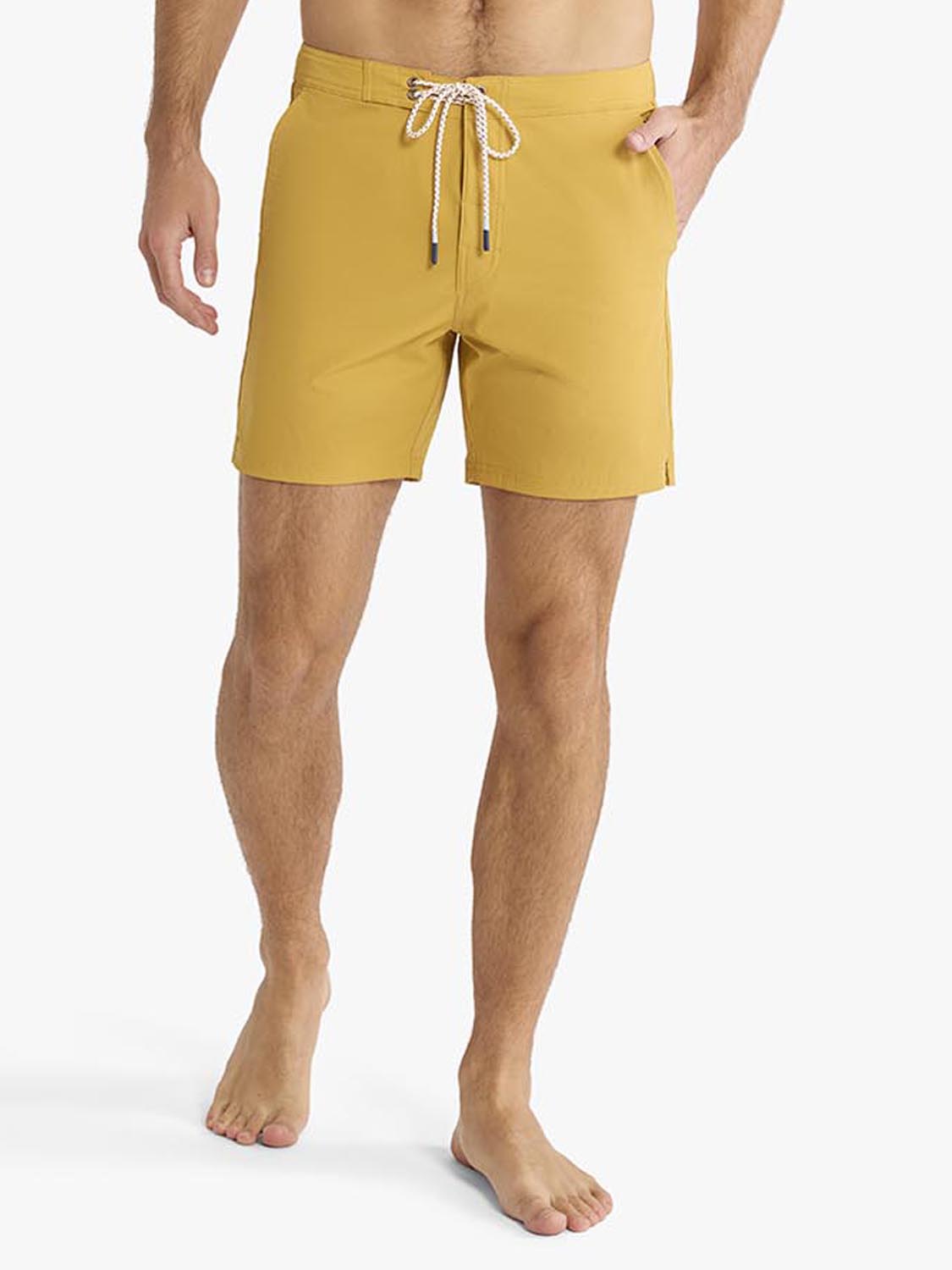 Man wearing gold board shorts with drawstring waistband and side pockets standing barefoot.