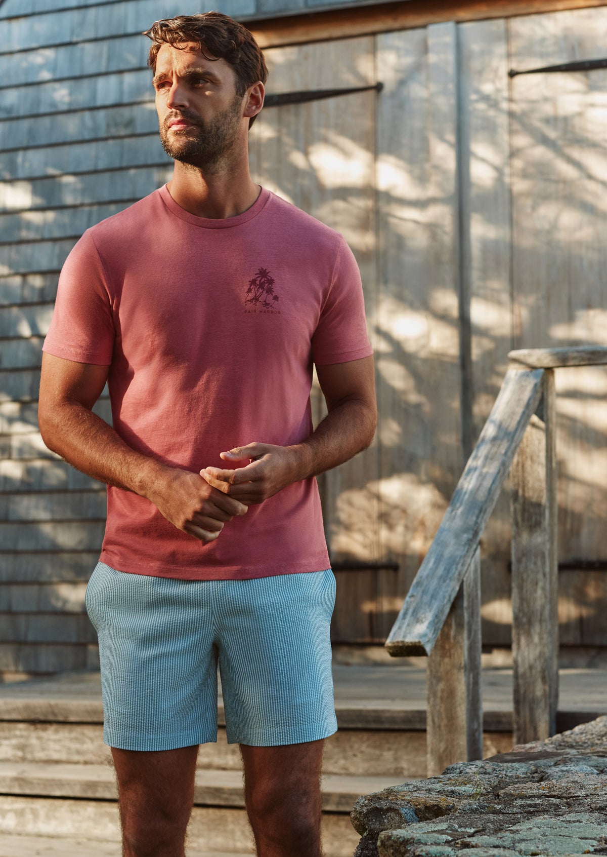Man wearing burgundy t-shirt and light blue seersucker shorts standing outdoors against weathered wooden structure.
