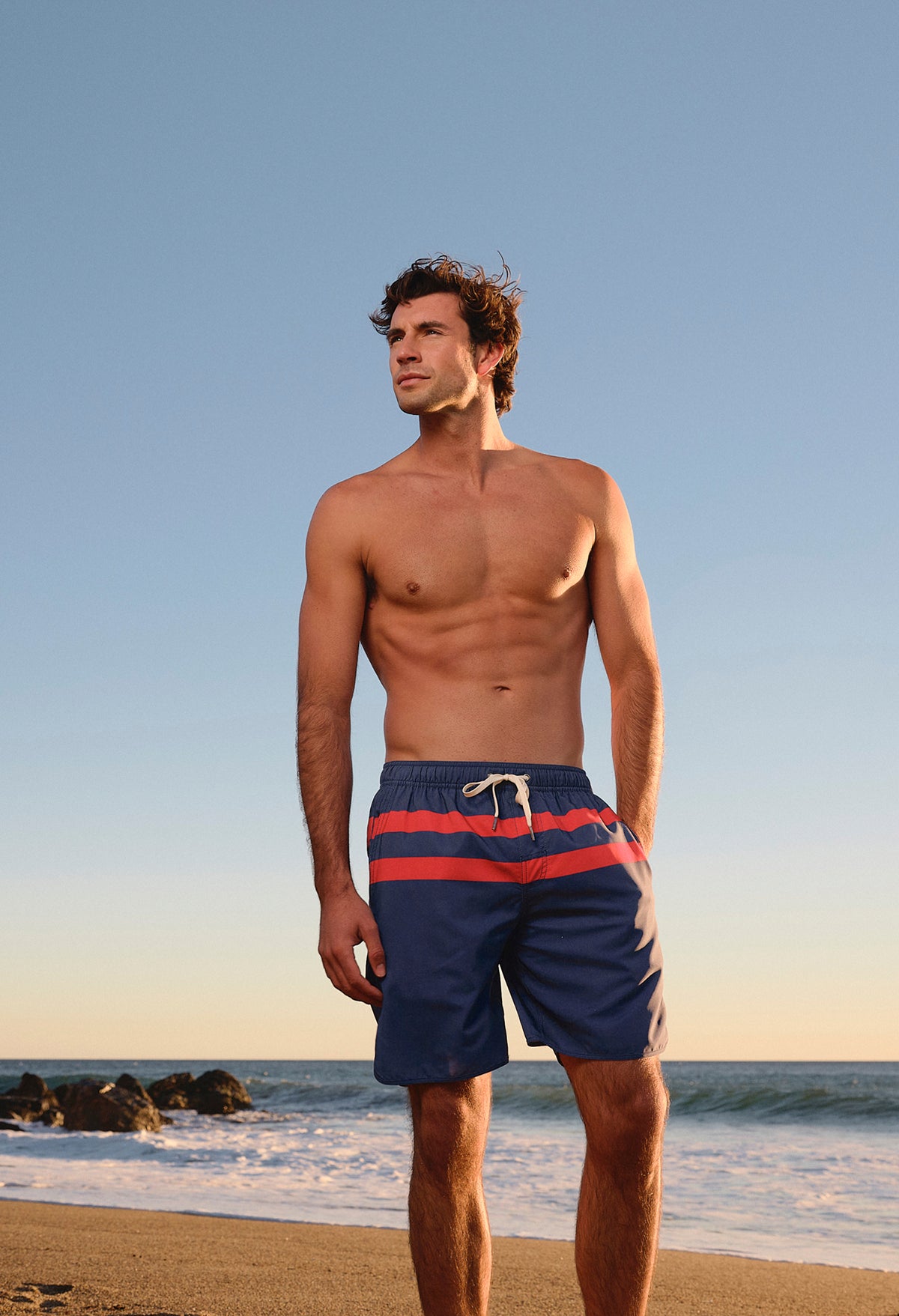 Man wearing navy swim shorts with red horizontal stripes standing on a beach.