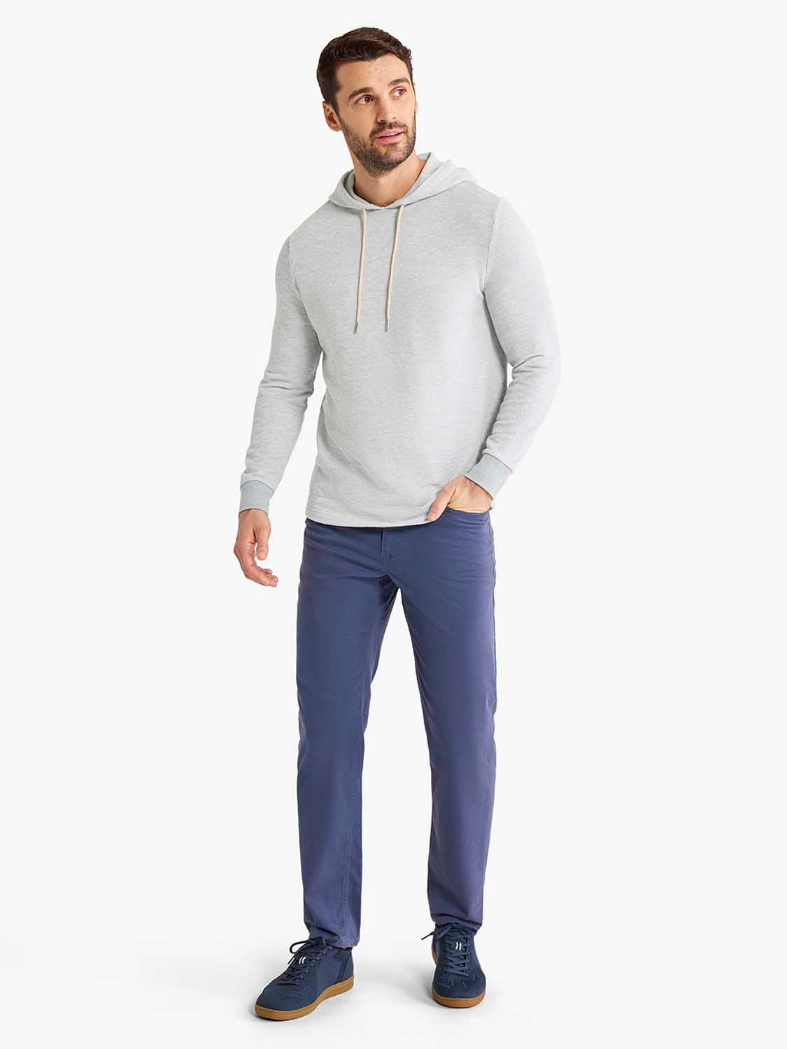 Man wearing heather grey hoodie with drawstring and navy blue pants standing against white background.