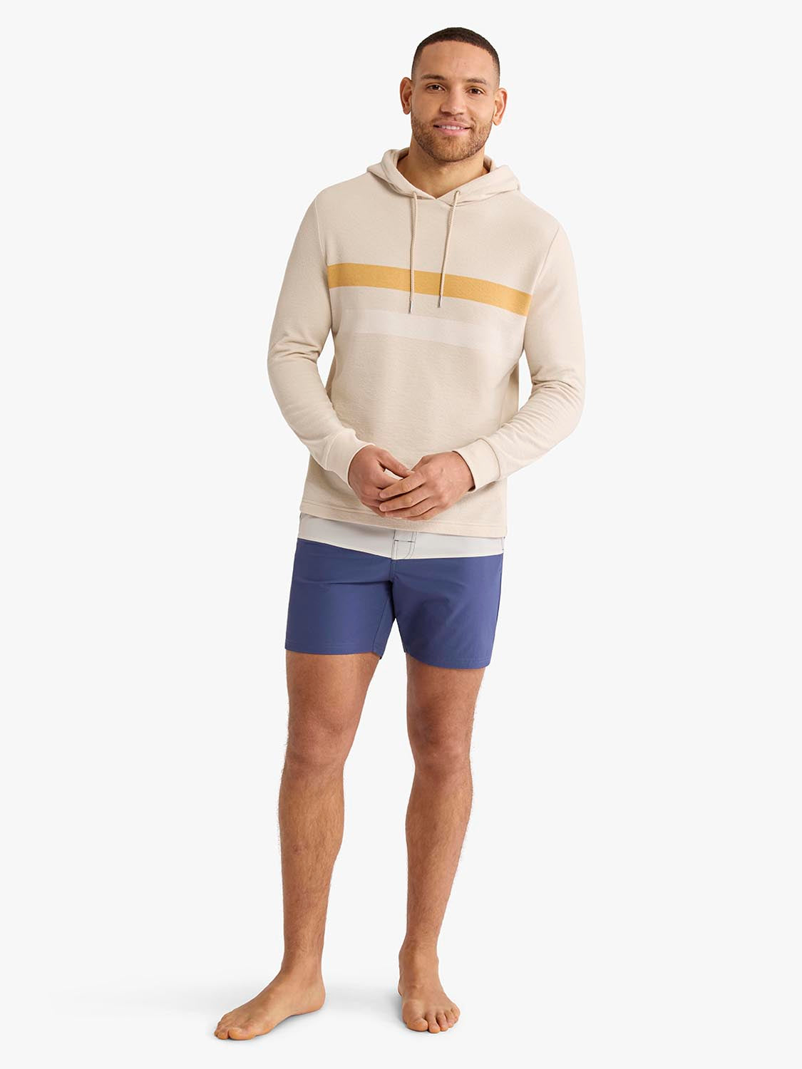 Man wearing a cream hoodie with a gold horizontal stripe across the chest and navy shorts.