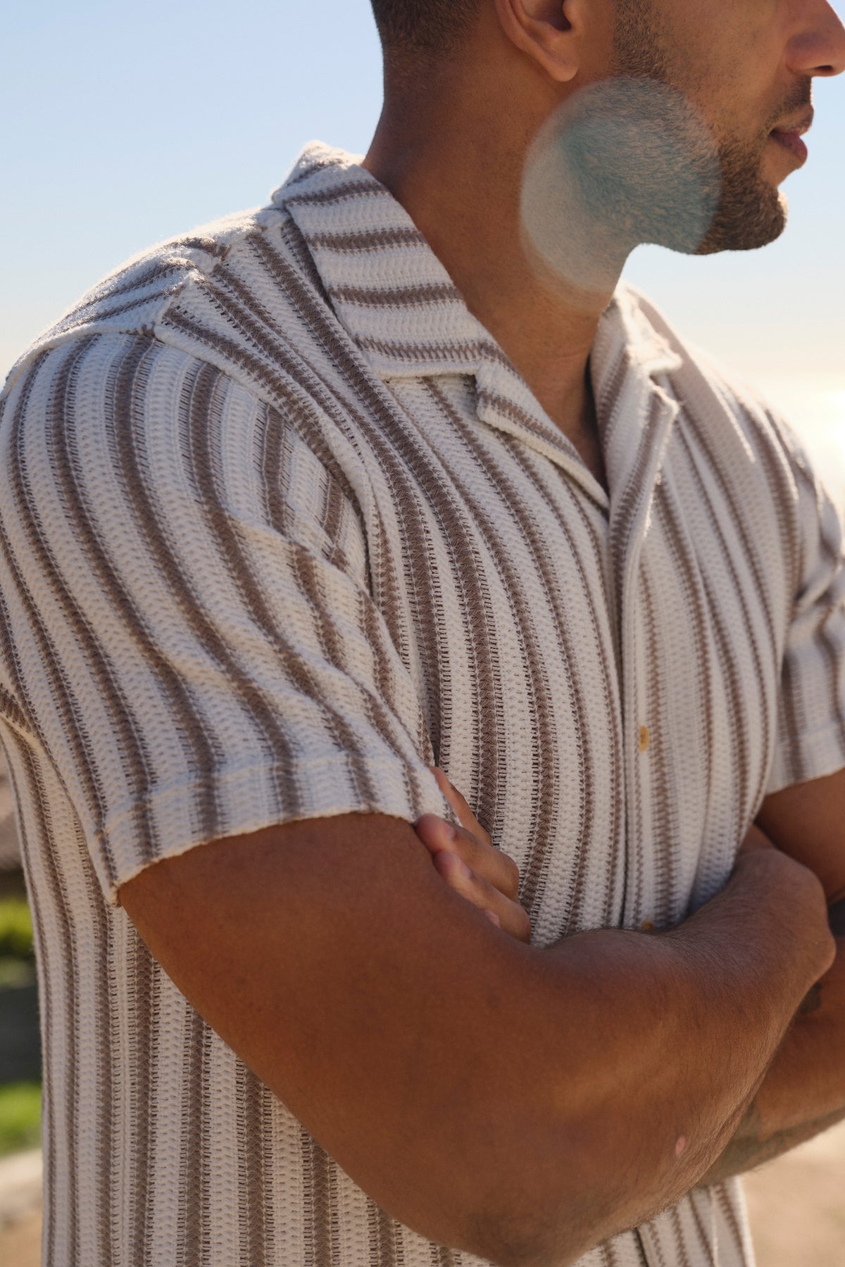 Man wearing a cream and taupe striped crochet short-sleeve shirt outdoors in natural daylight.