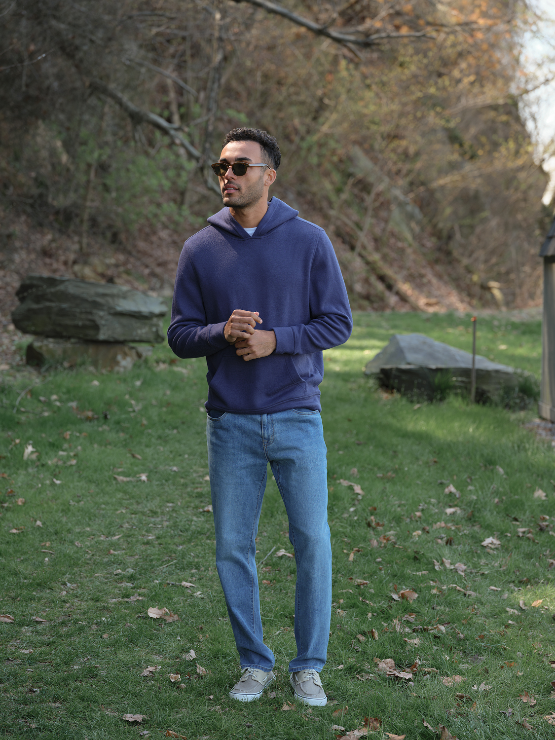 Man wearing a navy blue hoodie with blue jeans standing outdoors in a park setting.