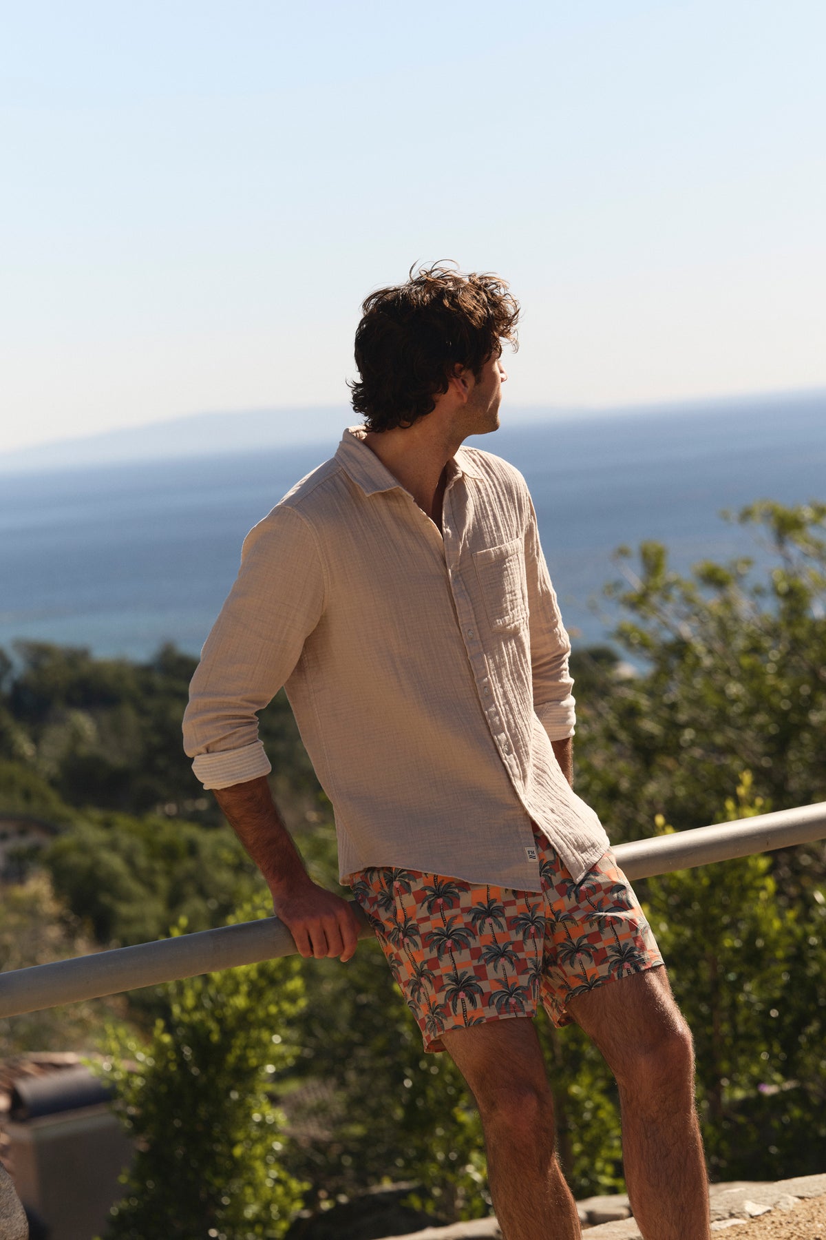 Man wearing beige shirt and geometric patterned swim shorts stands on an overlook facing the ocean.