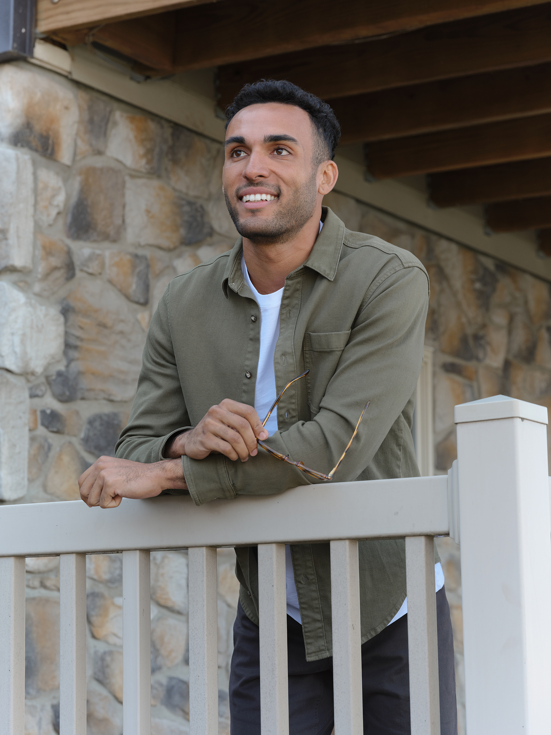 Man wearing olive denim shirt over white shirt leaning on porch railing outdoors.