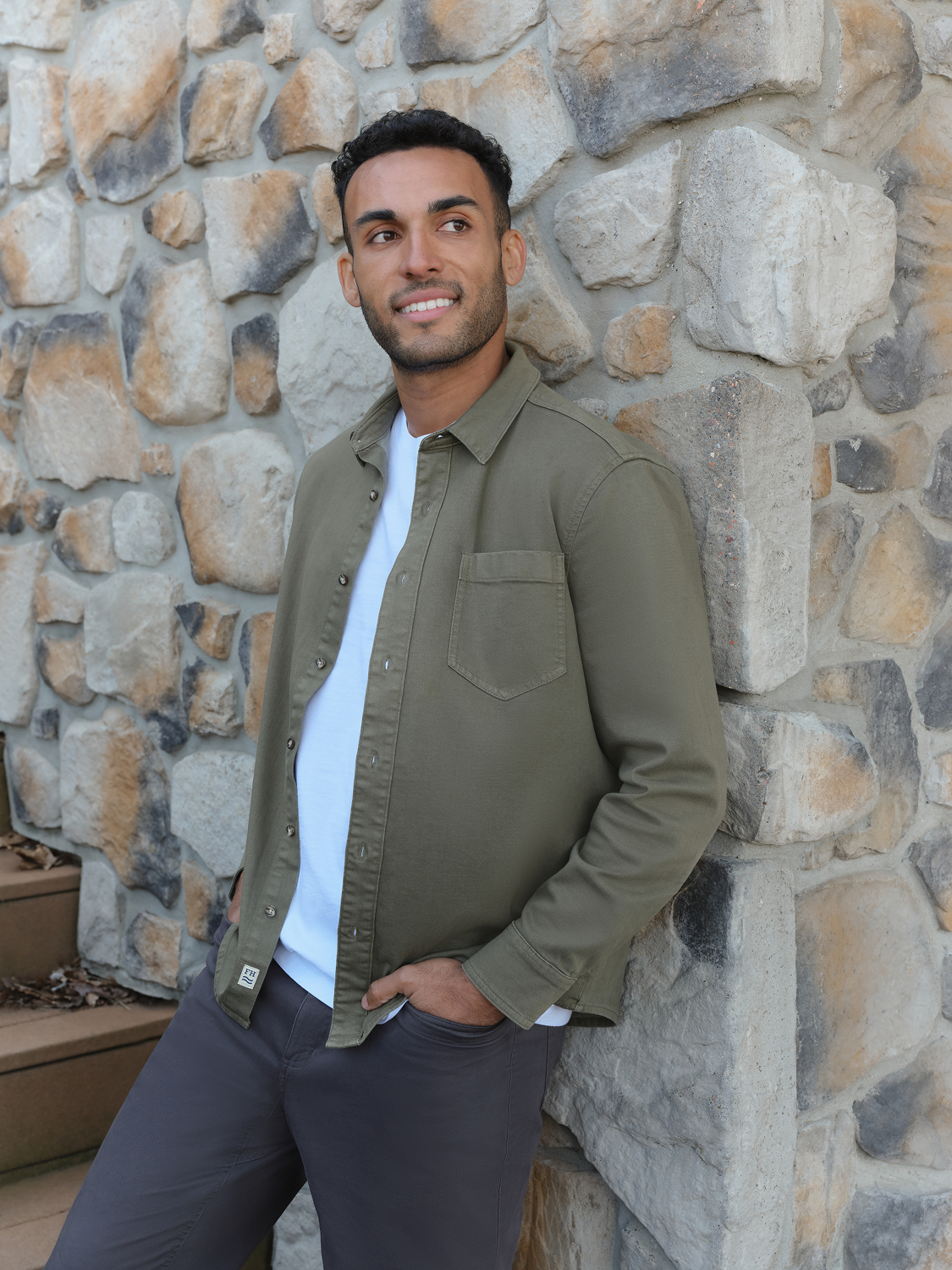 Man wearing an olive-colored denim shirt with a white t-shirt underneath against a stone wall.