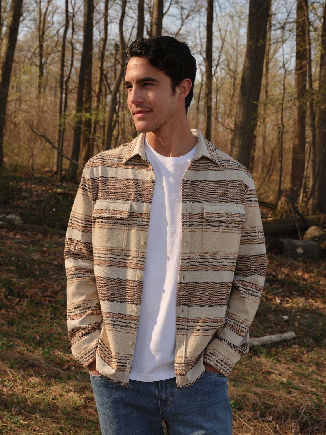 Man wearing a tan and brown striped flannel shirt over a white t-shirt and blue jeans.