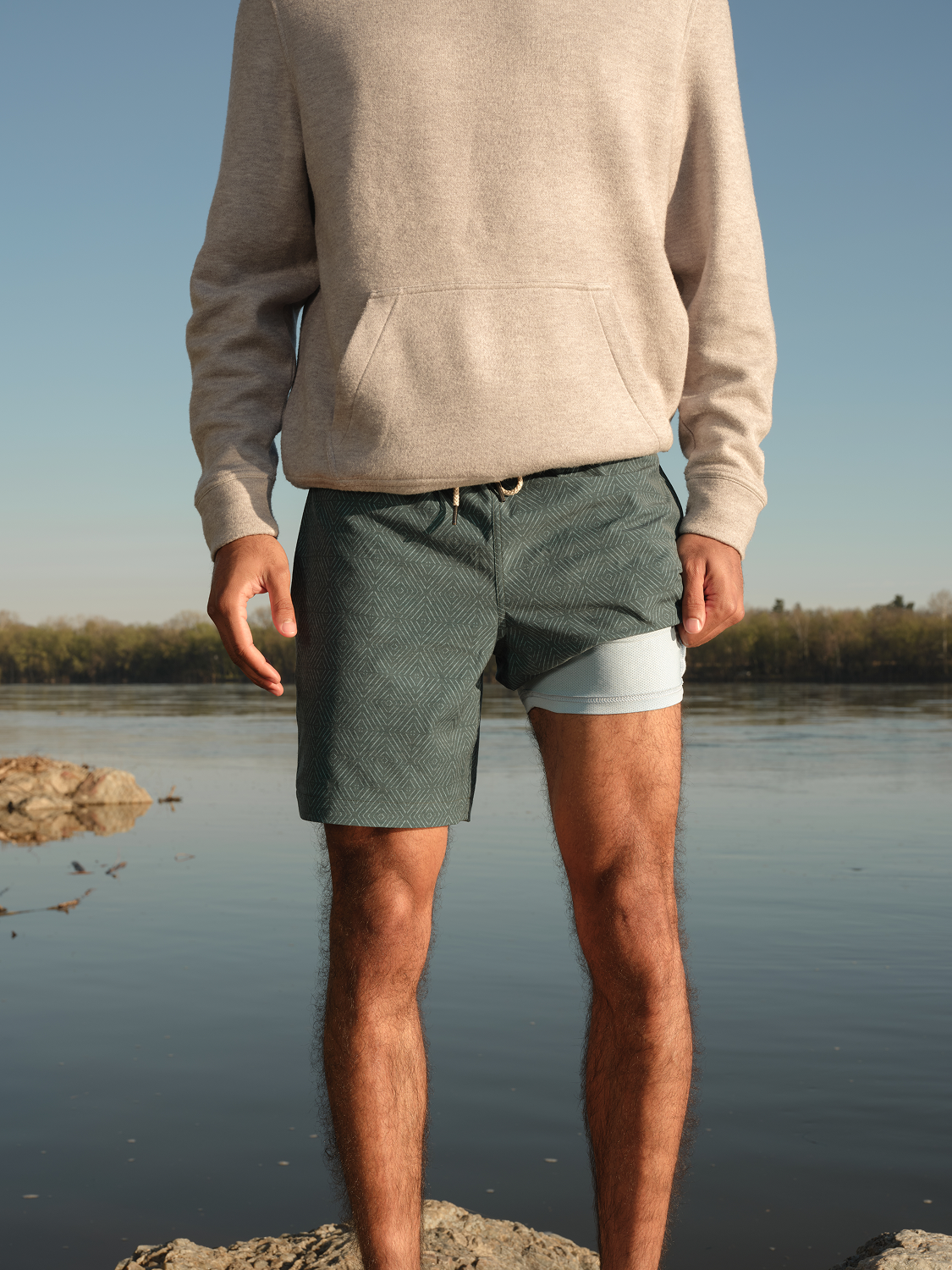 Man wearing green shibori-patterned swim trunk with white liner detail standing by water.