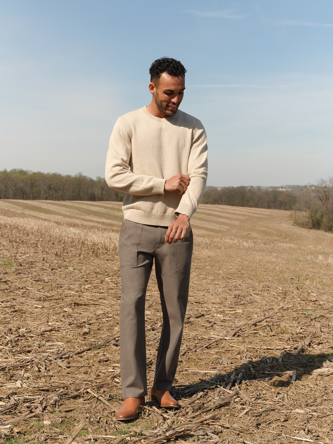 Man wearing a cream-colored crew neck sweater and gray pants standing in an open field.
