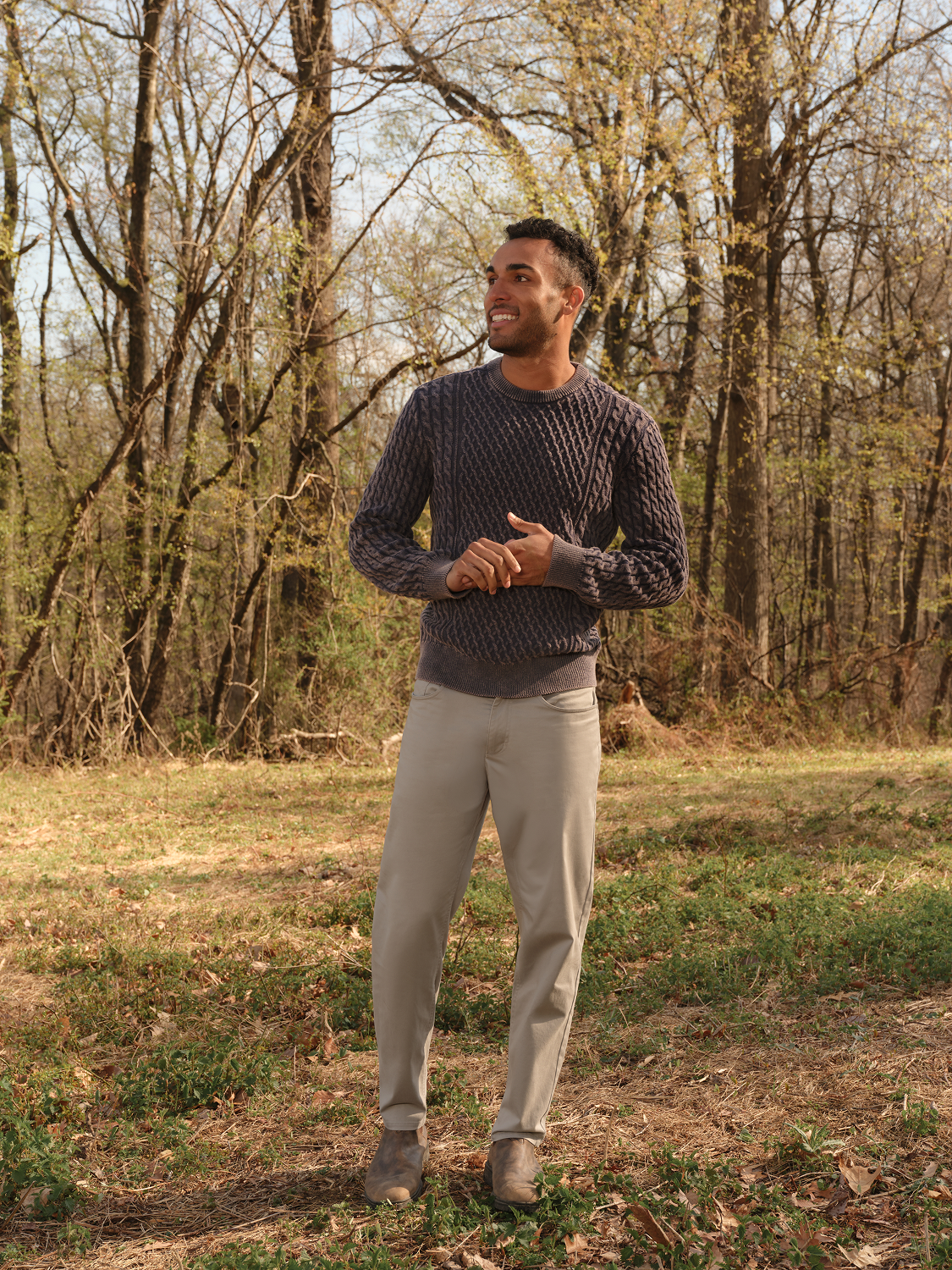 Man wearing an oak-colored cable knit sweater and beige pants standing outdoors among bare trees.