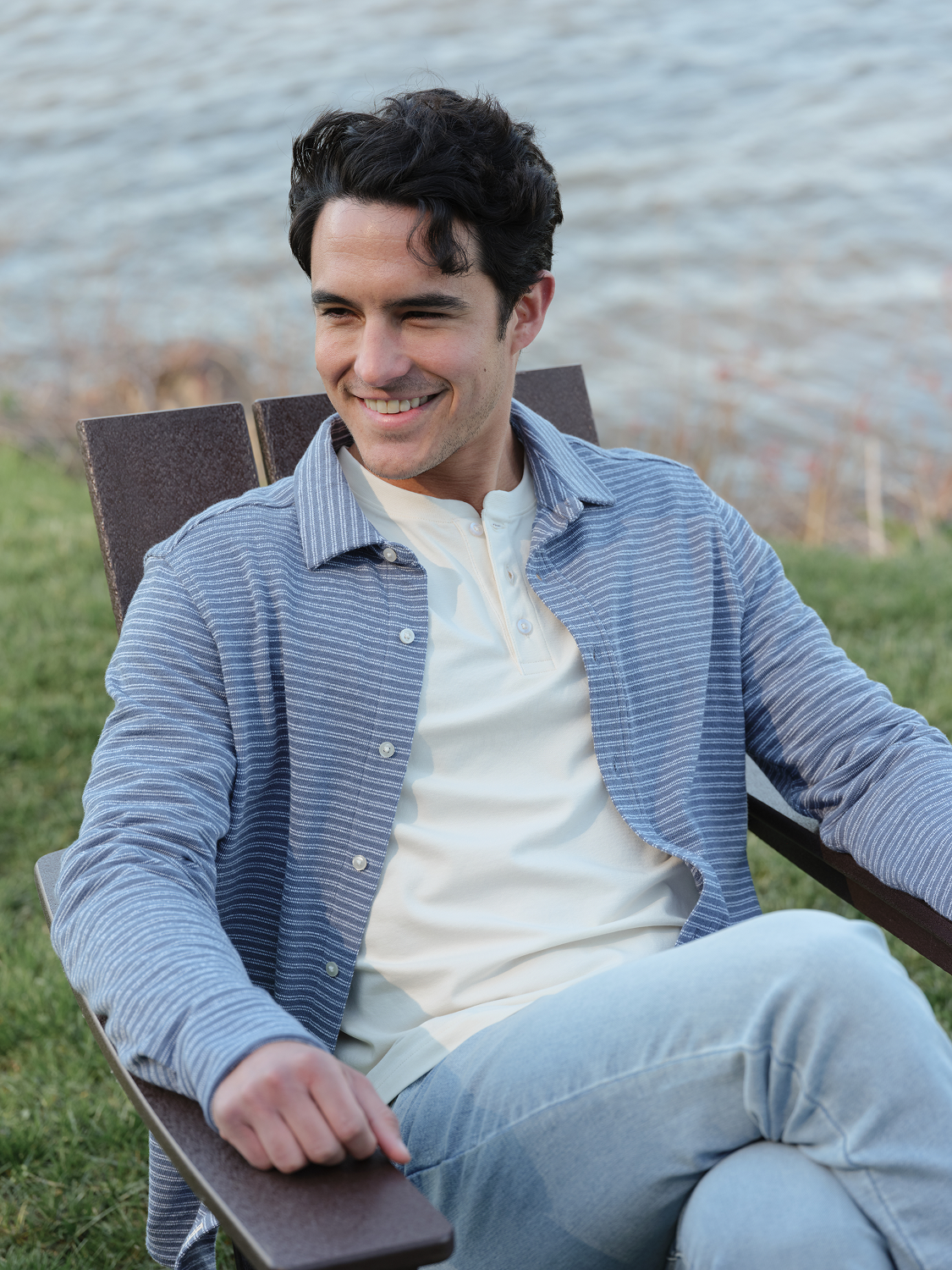 Man wearing a blue striped button-up shirt over a cream henley sitting on a bench by water.