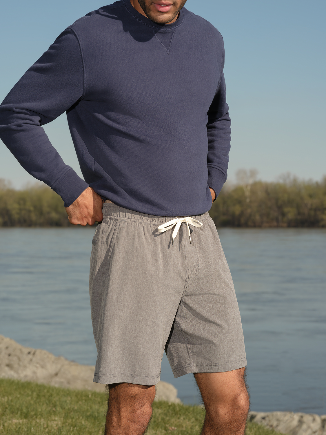 Man wearing a dark navy crewneck sweatshirt and gray shorts with drawstring standing by water.