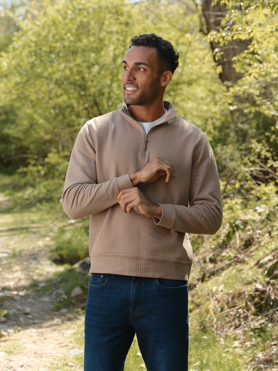 Man wearing a beige quarter zip pullover and dark jeans standing outdoors surrounded by green foliage.